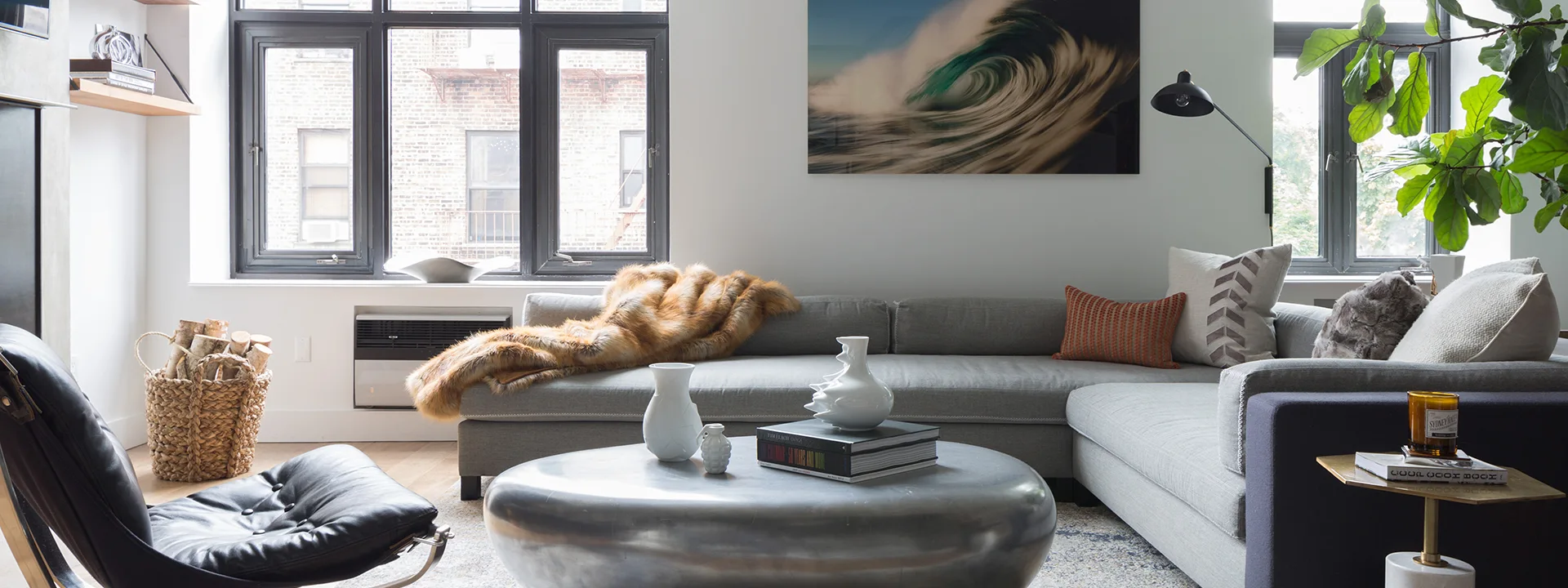 A light-filled living room features a grey sectional sofa, a round metallic coffee table, black-framed windows, and a black leather lounge chair.