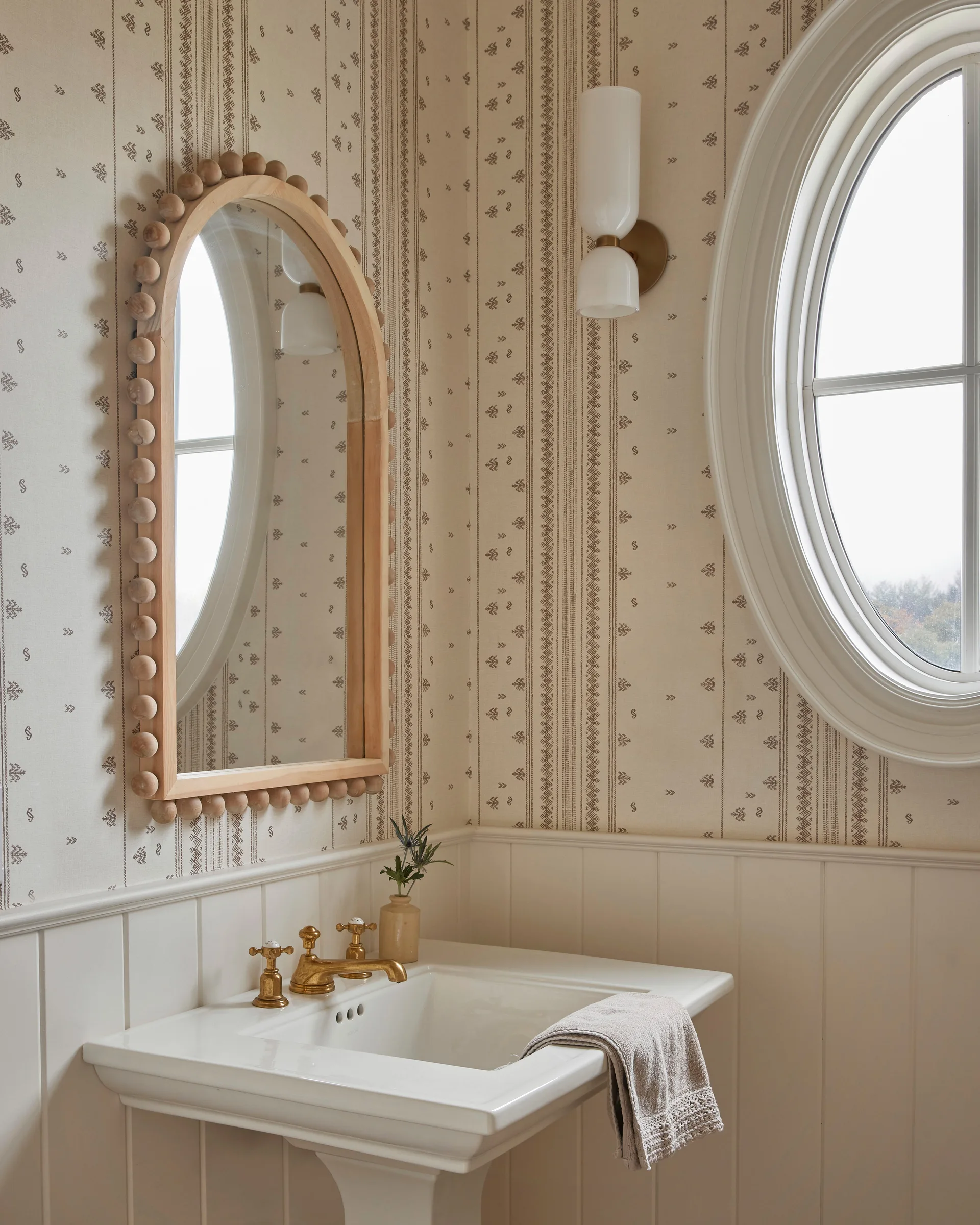 A classic powder room features a white pedestal sink, a wood-framed arched mirror with bead detailing, patterned wallpaper, and beadboard wainscoting.