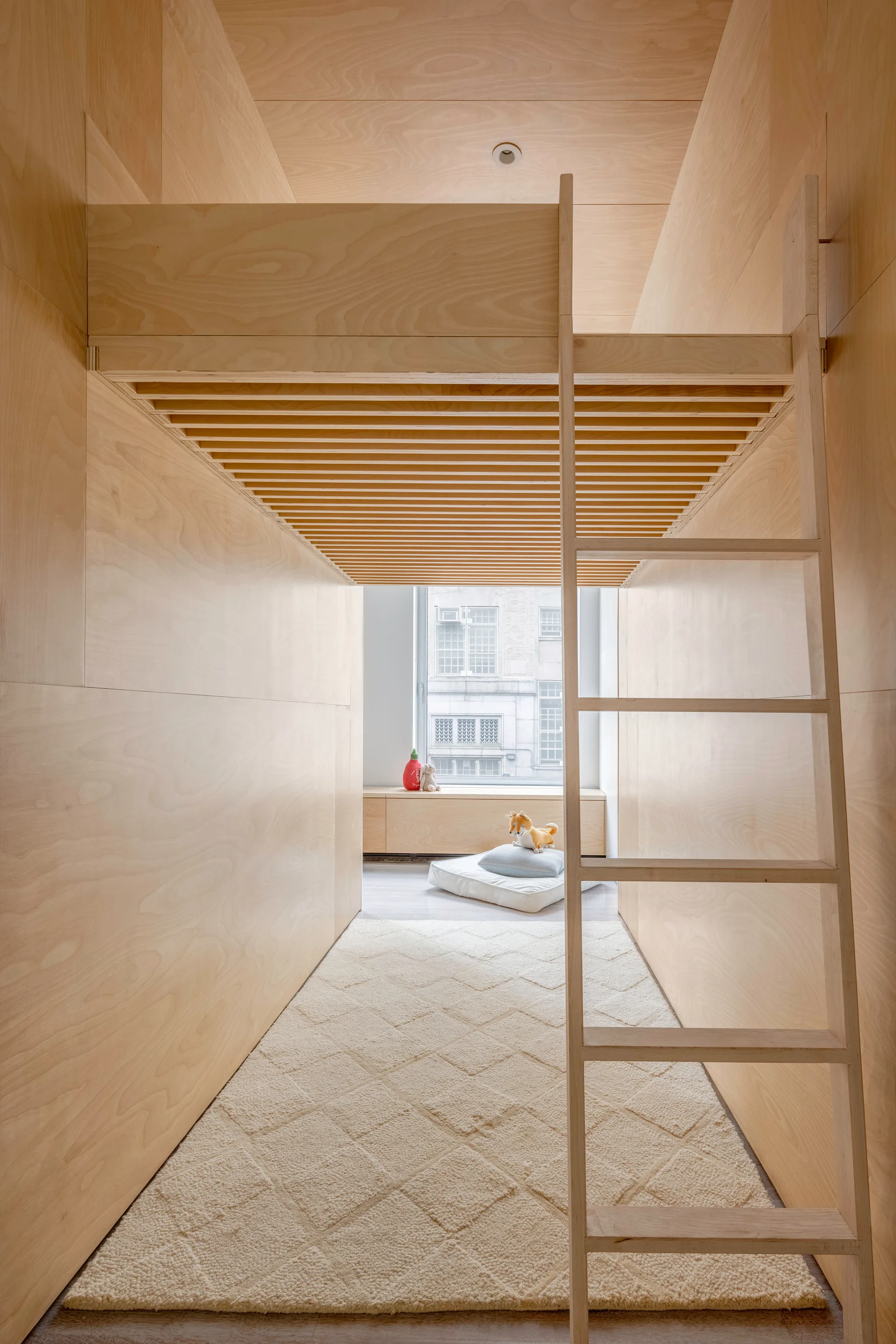 A small bedroom wrapped in light plywood paneling features a lofted bed with a wooden ladder, a diamond-patterned rug, and a window seat.