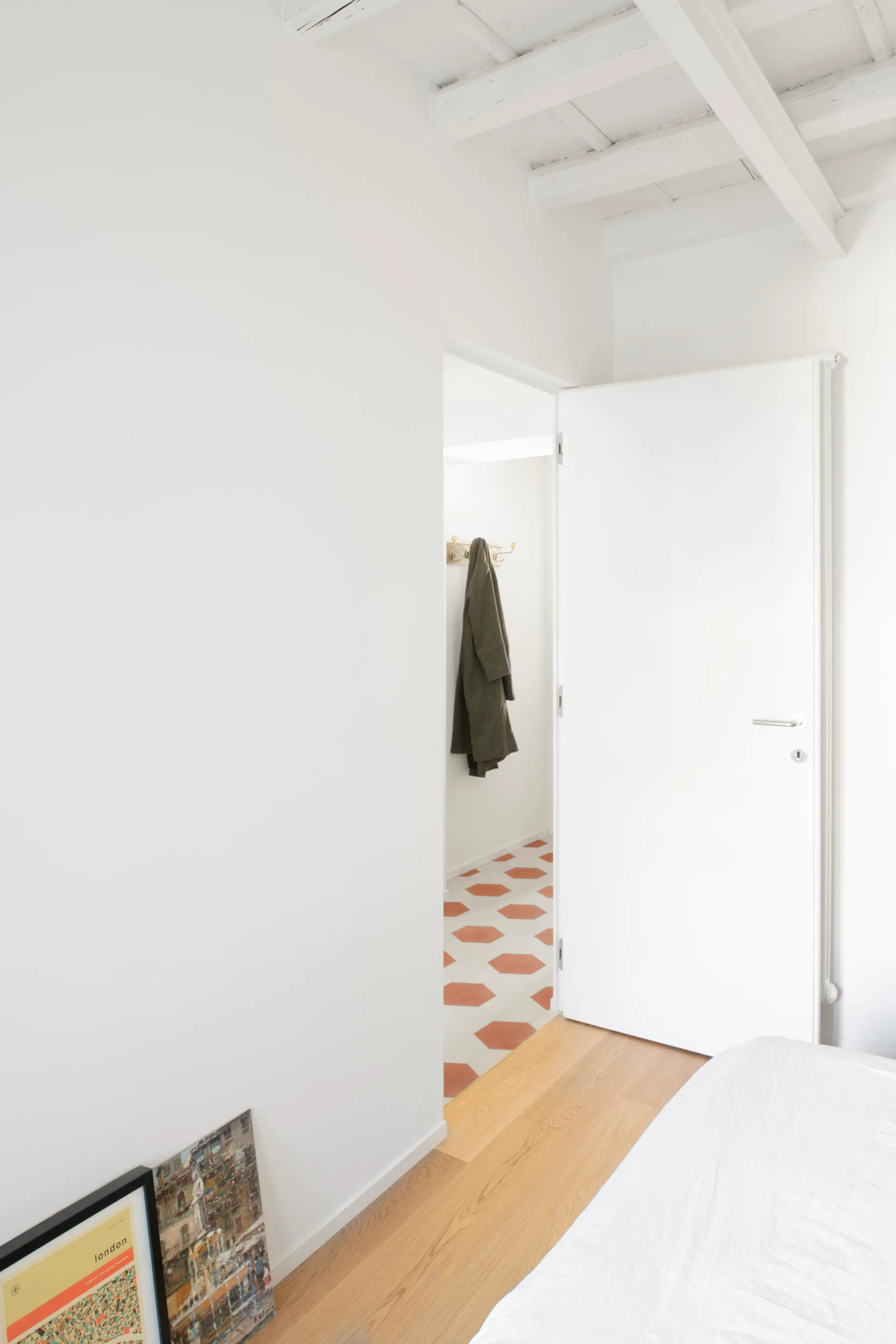 An open white door reveals a hallway with terracotta and white hexagonal floor tiles, a hanging green coat, and exposed ceiling beams.