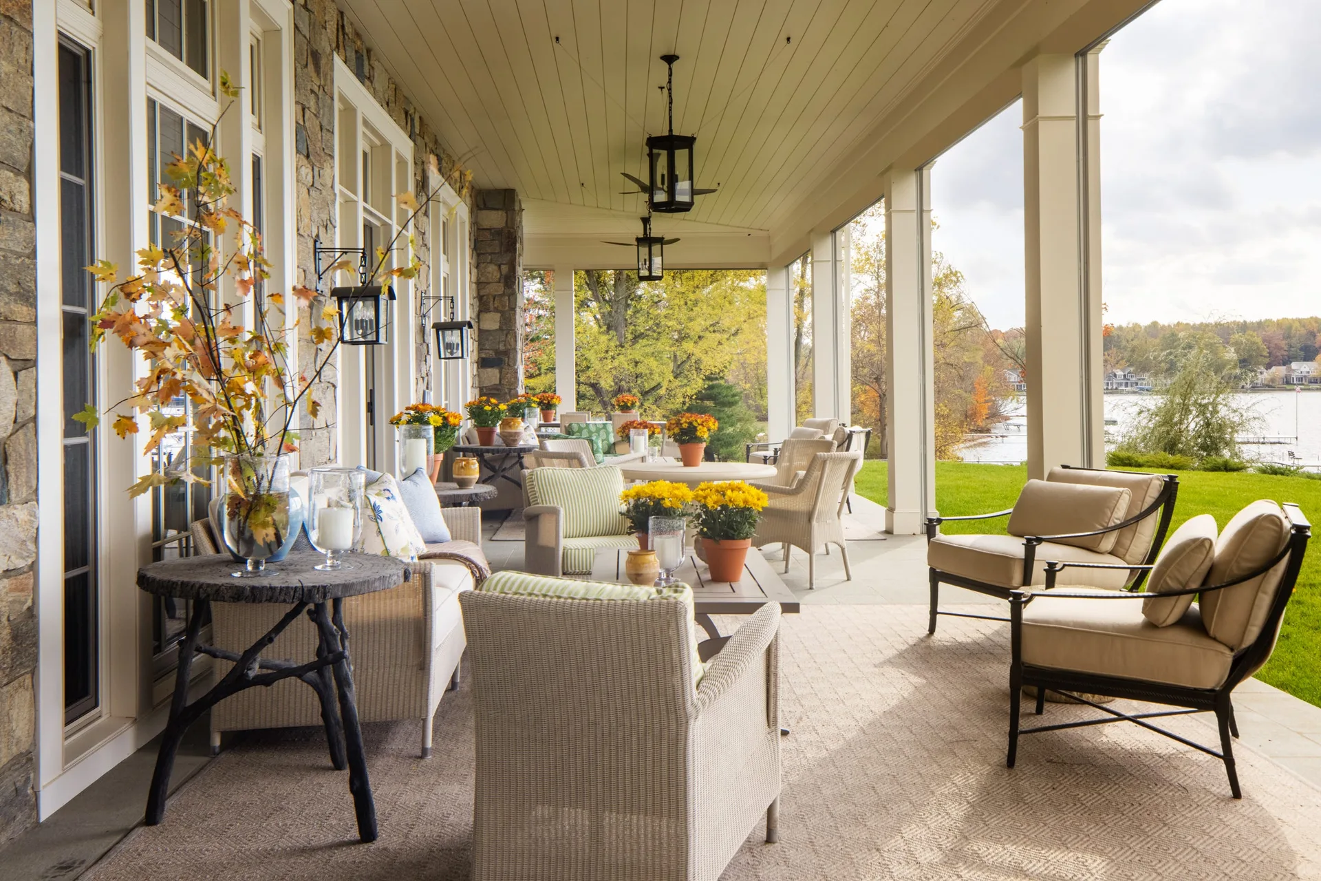 A covered stone patio overlooking a lake features neutral-colored wicker sofas and chairs, black hanging lanterns, and autumn flowers.