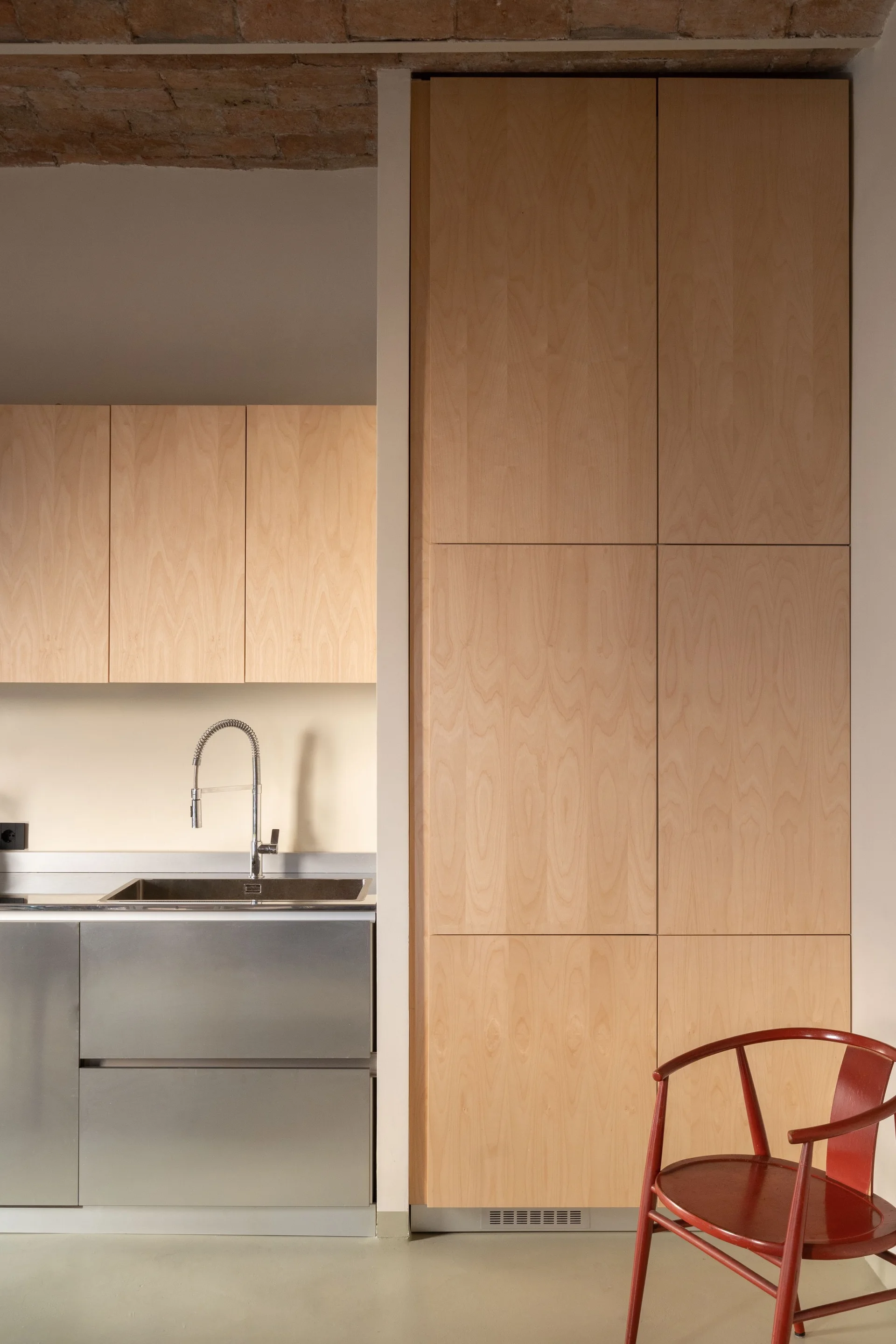 A minimalist kitchen featuring tall light wood cabinets, stainless steel lower drawers, a chrome faucet, and a red wooden armchair.