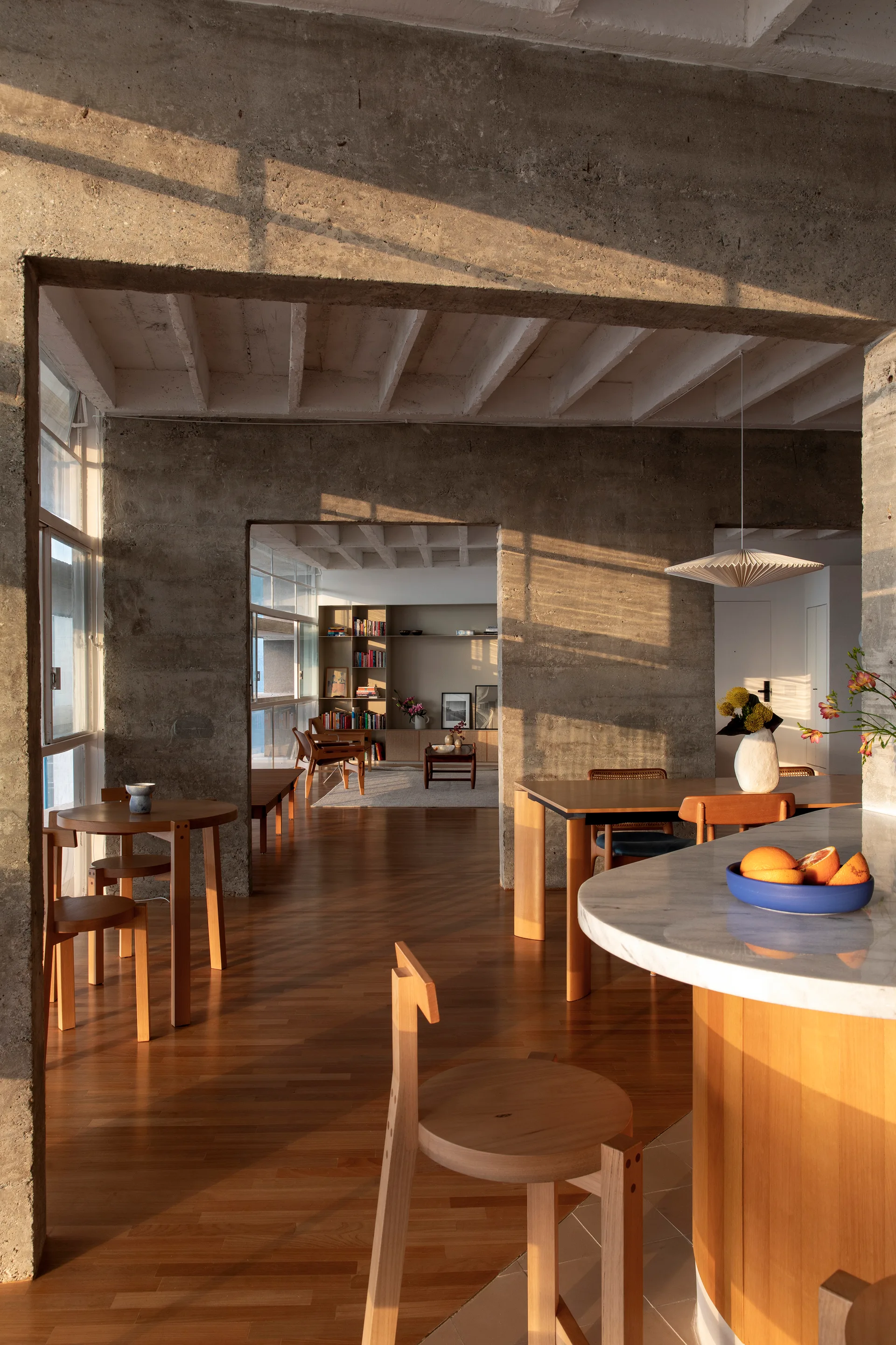 An open dining area features raw concrete walls, warm wood flooring, mid-century modern chairs, and sunlight streaming through large windows.
