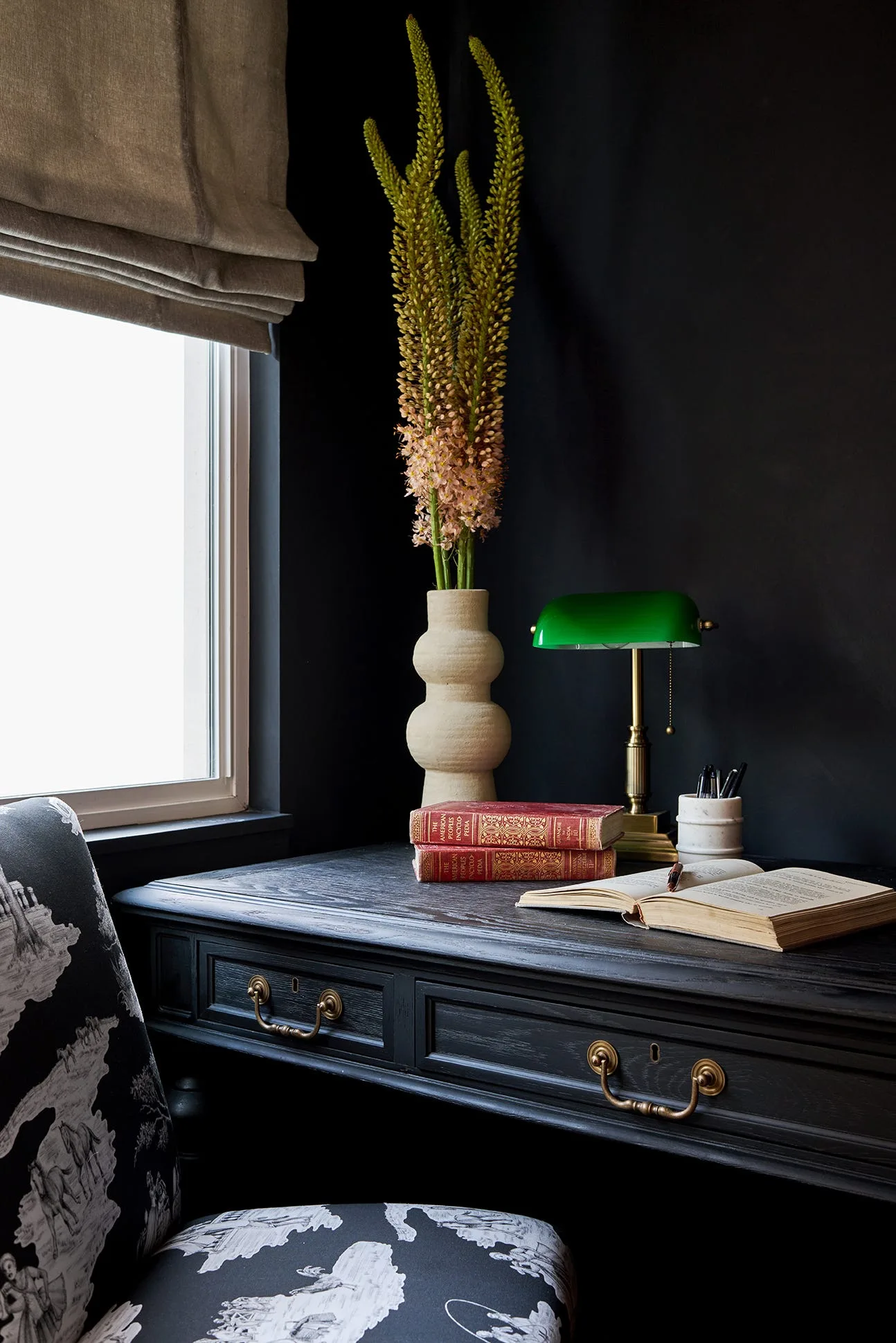 A close-up of a black wooden desk with brass hardware featuring an open book, vintage books, a green banker lamp, and a tall flower vase next to a toile chair.