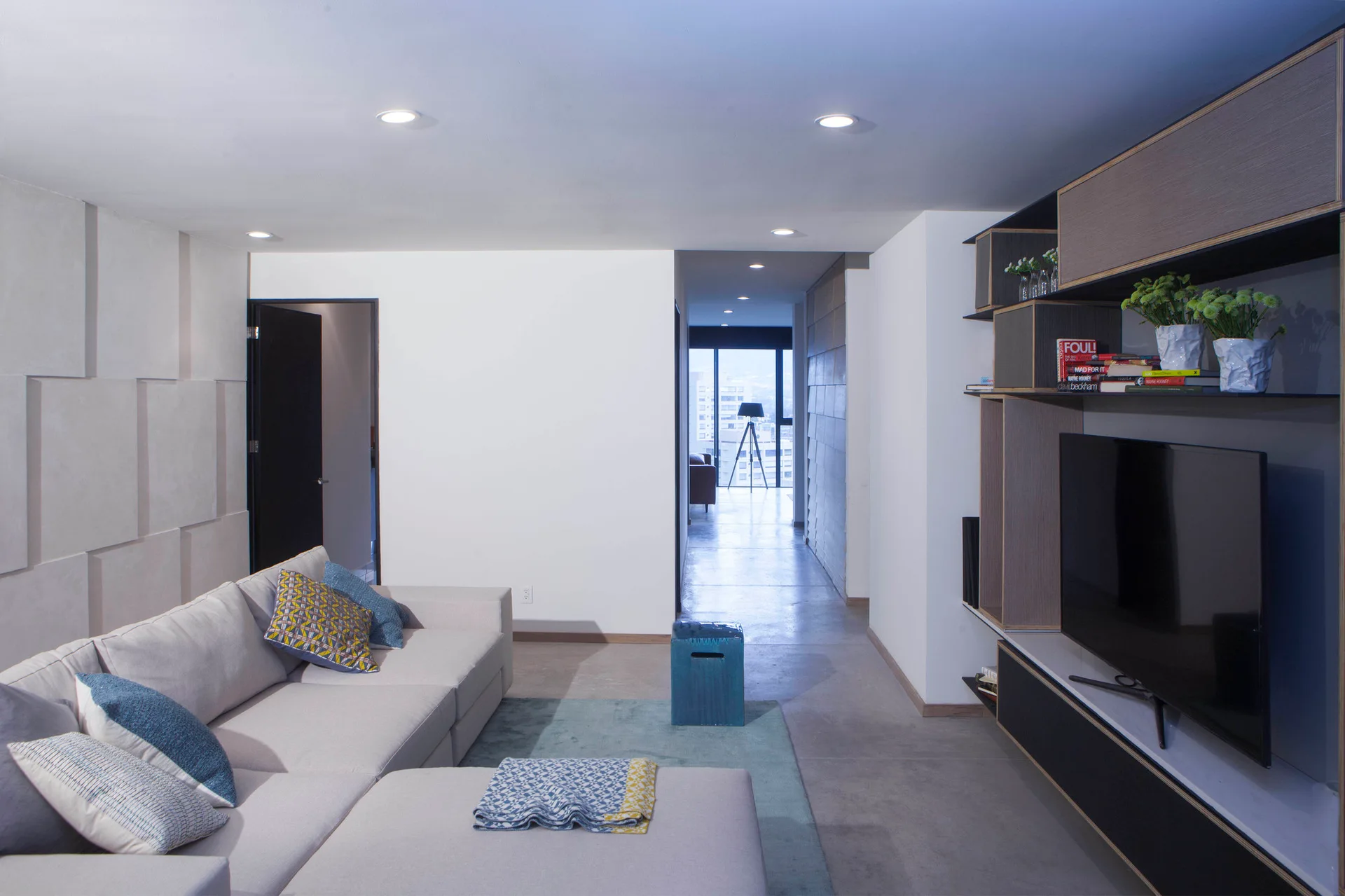 A light gray sectional sofa and a built-in media console sit on a gray concrete floor with a blue rug and textured white wall panels.