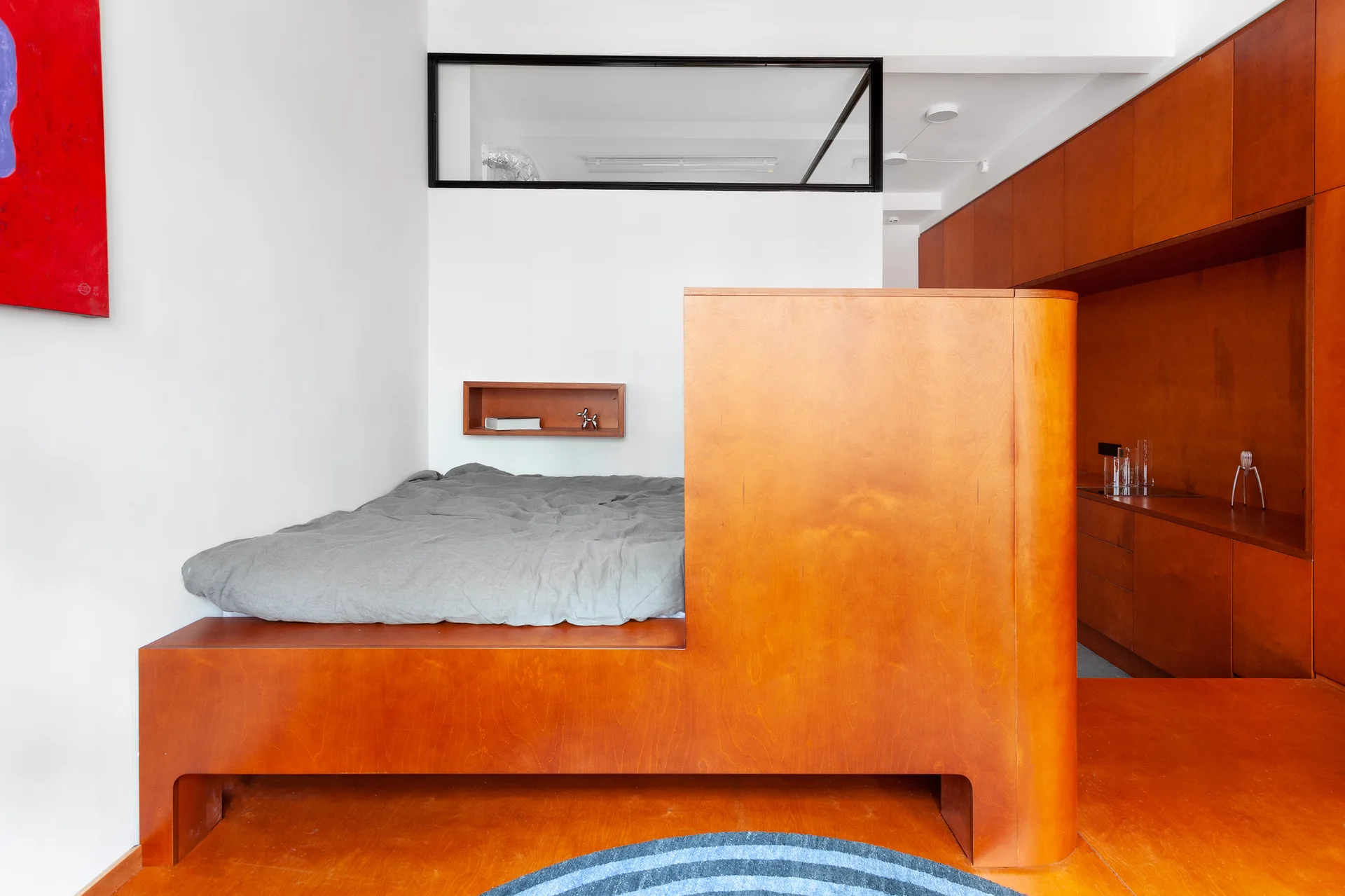 A custom warm wood bed with grey bedding is integrated with a built-in partition wall next to matching wood cabinetry.