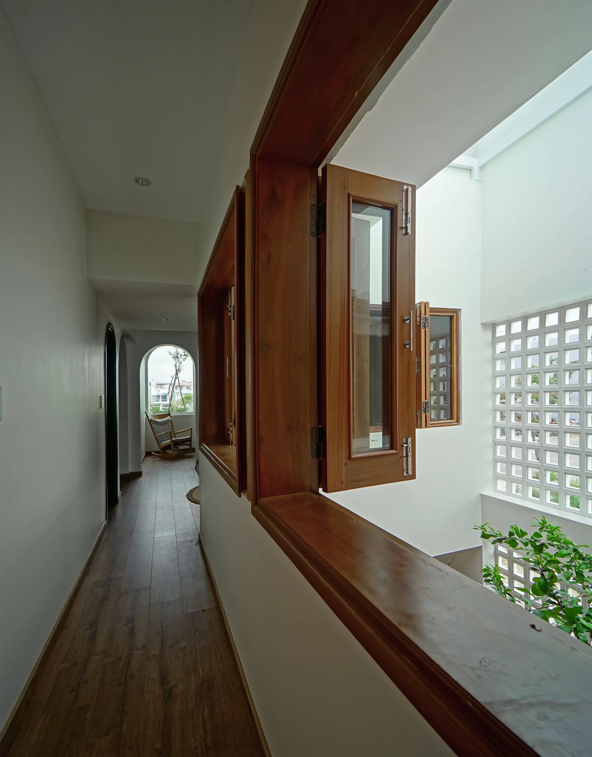 A narrow white hallway features wood plank floors, deep wooden casement windows, and a view of an atrium with a geometric breeze block wall.