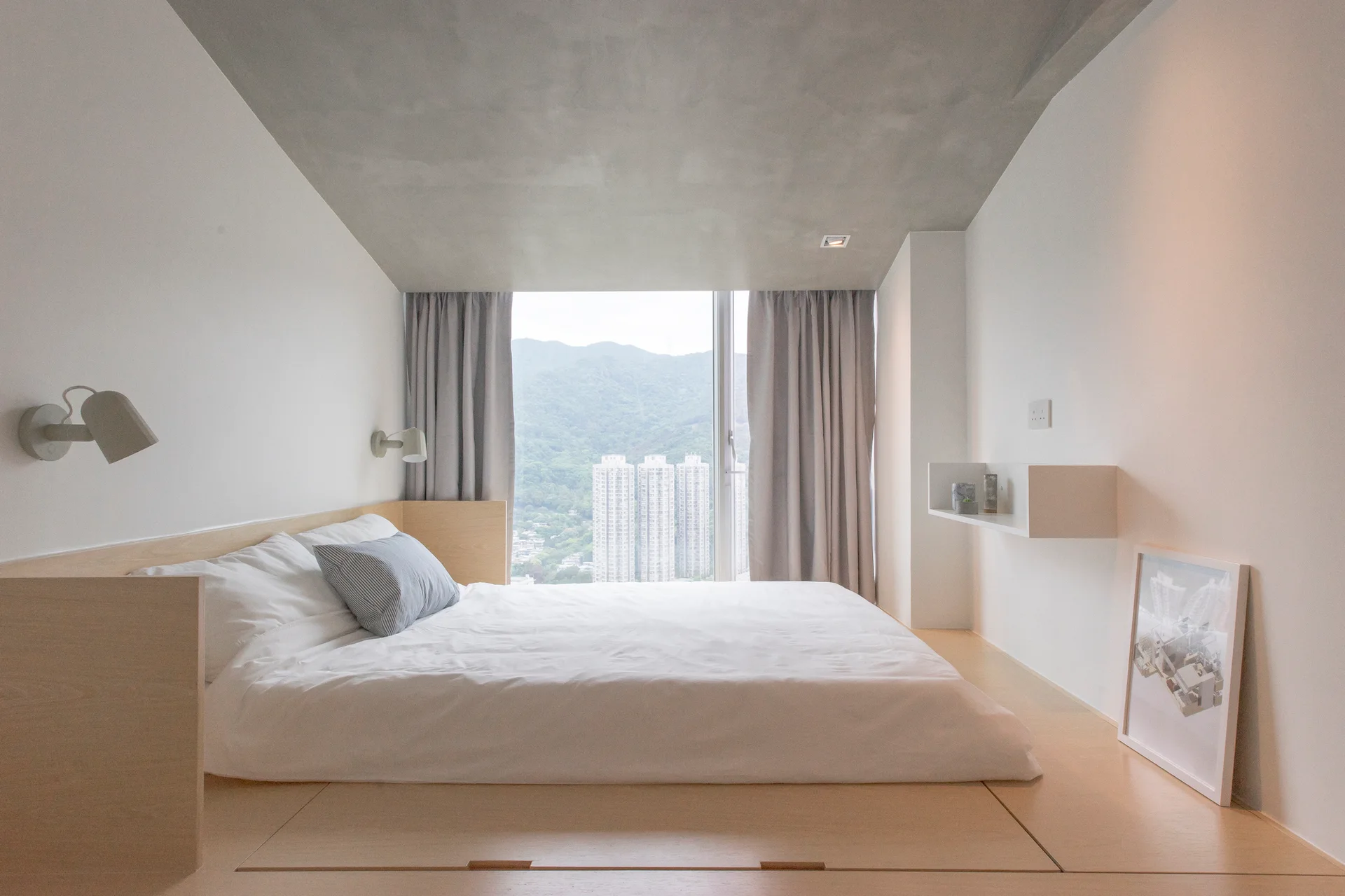 A minimalist bedroom features a low platform bed integrated into a light wood base, white walls, and a smooth grey concrete ceiling.
