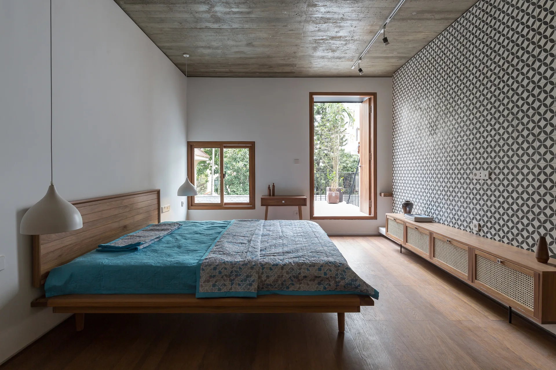 A modern bedroom features a wooden platform bed, a patterned geometric accent wall, and an exposed raw concrete ceiling.