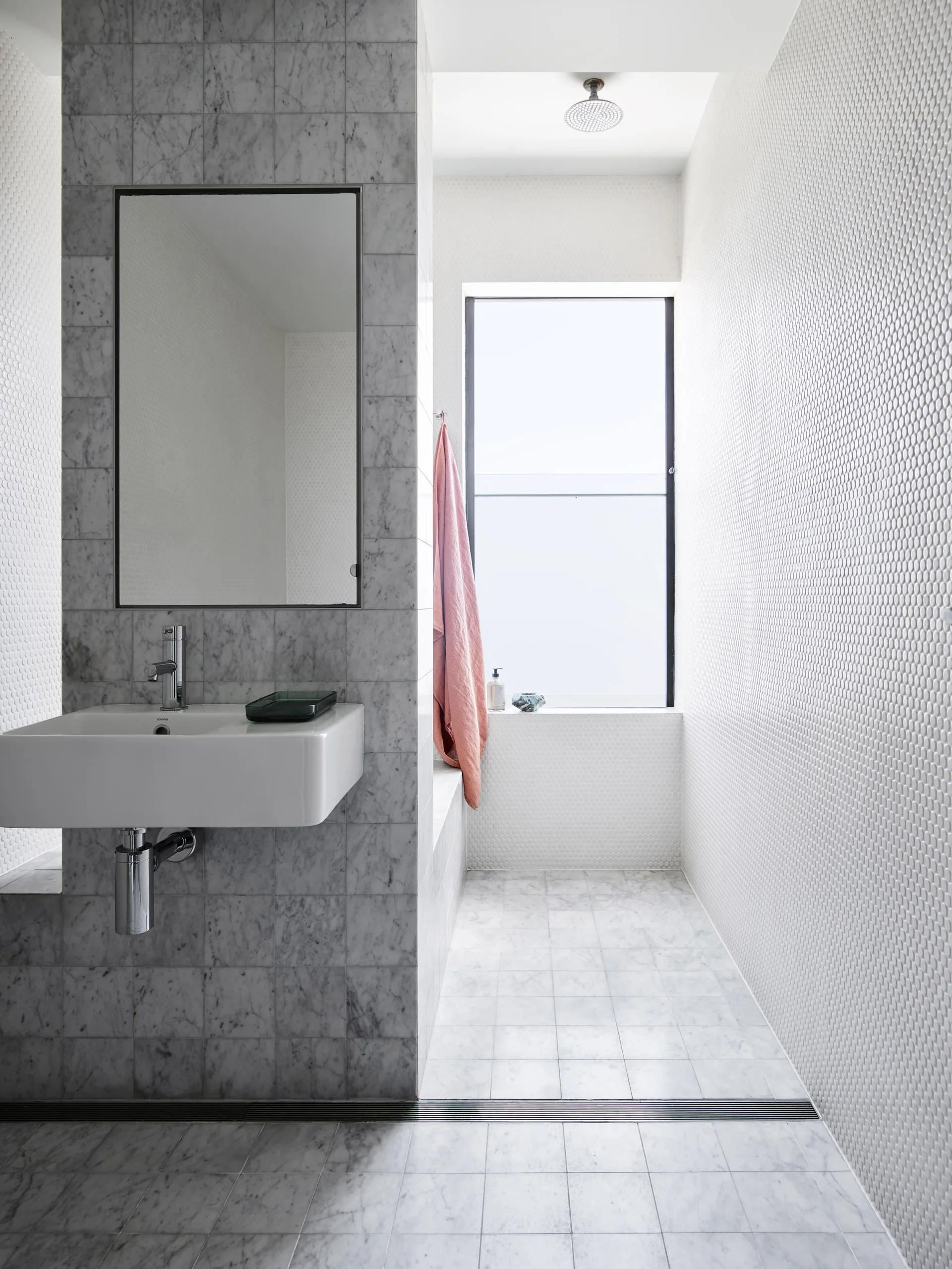 A modern bathroom showing a wall-mounted sink on a marble partition, surrounded by white penny-tiled walls and a wet-room shower area by a window.