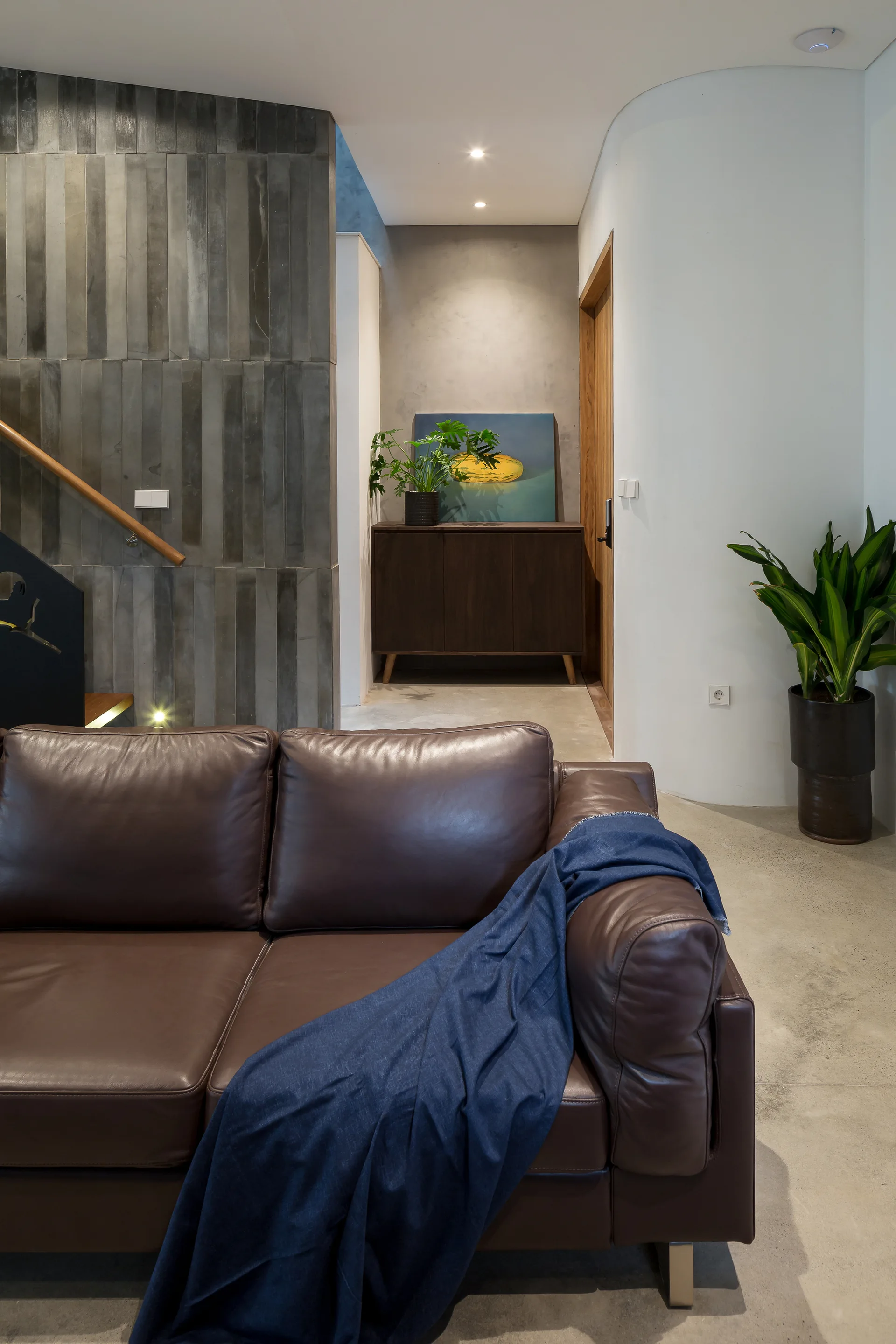 A modern living space featuring a brown leather sofa, vertical dark grey wall tiles, and a curved white wall opening into a hallway.
