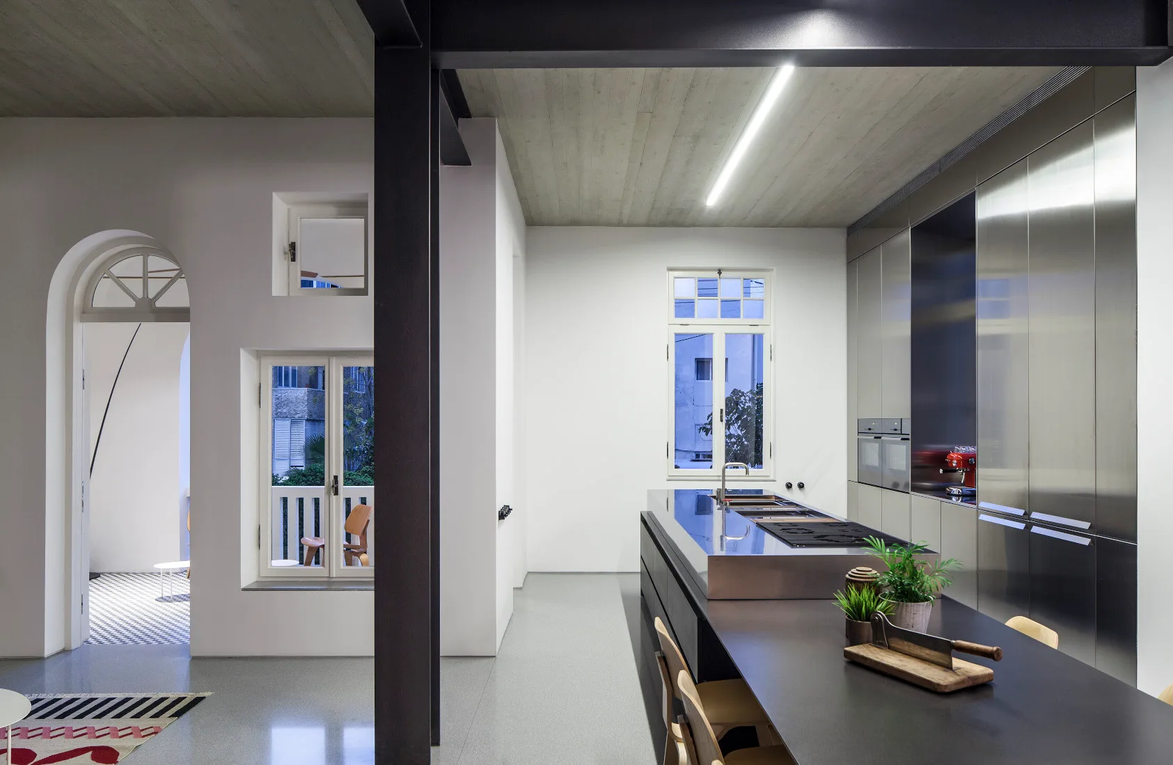 A minimalist kitchen features a large stainless steel island, full-height metal cabinets, a textured concrete ceiling, and a dark structural column.