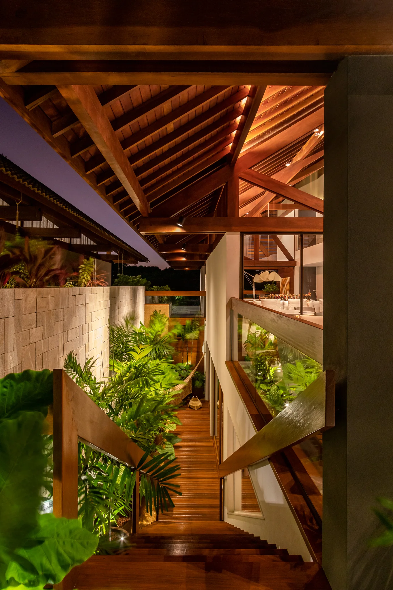A wooden staircase descends next to a lush tropical courtyard with a stone wall under an exposed timber roof.