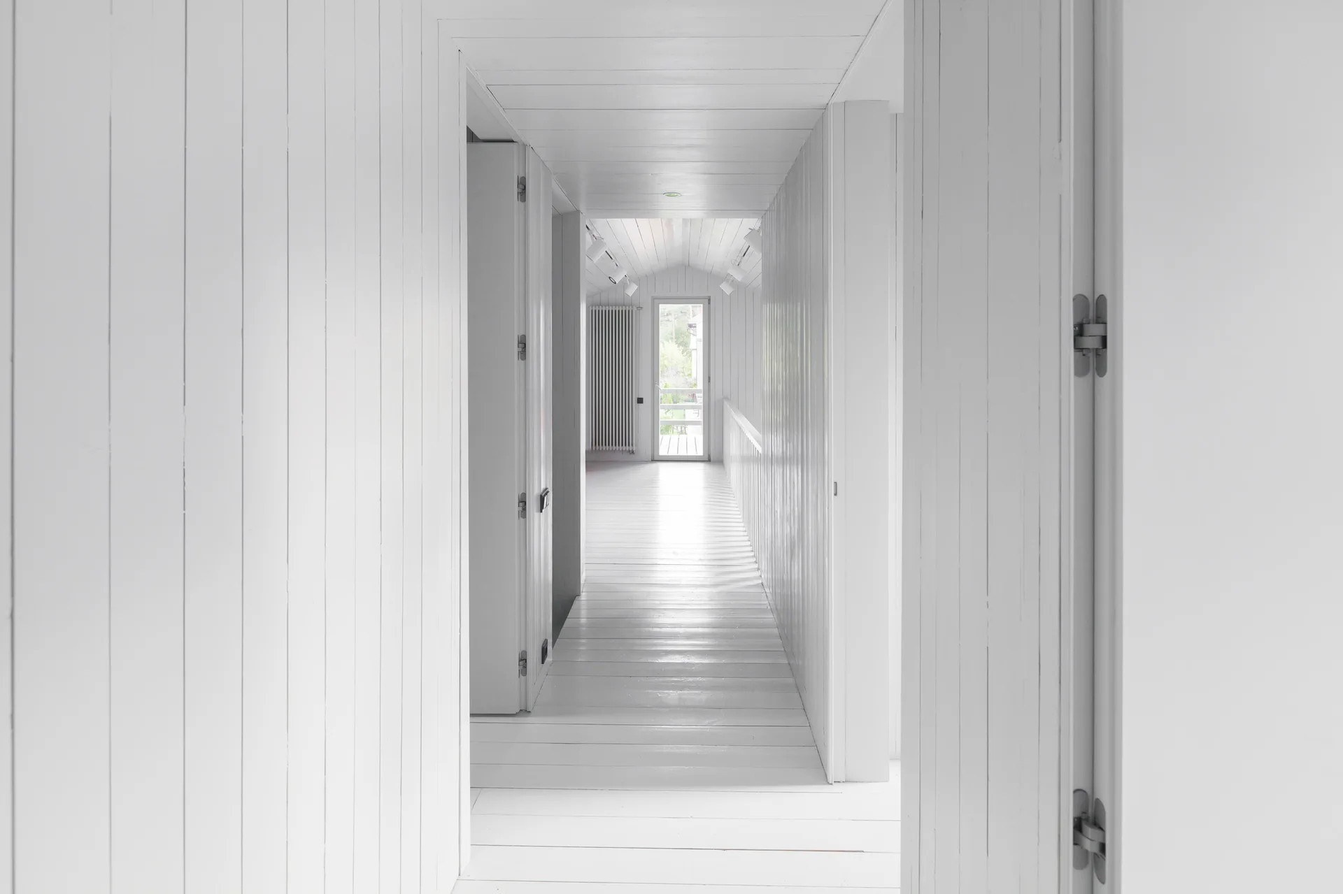 A long, bright hallway features white painted vertical wood paneling on the walls and matching white painted wood floorboards leading to a window.