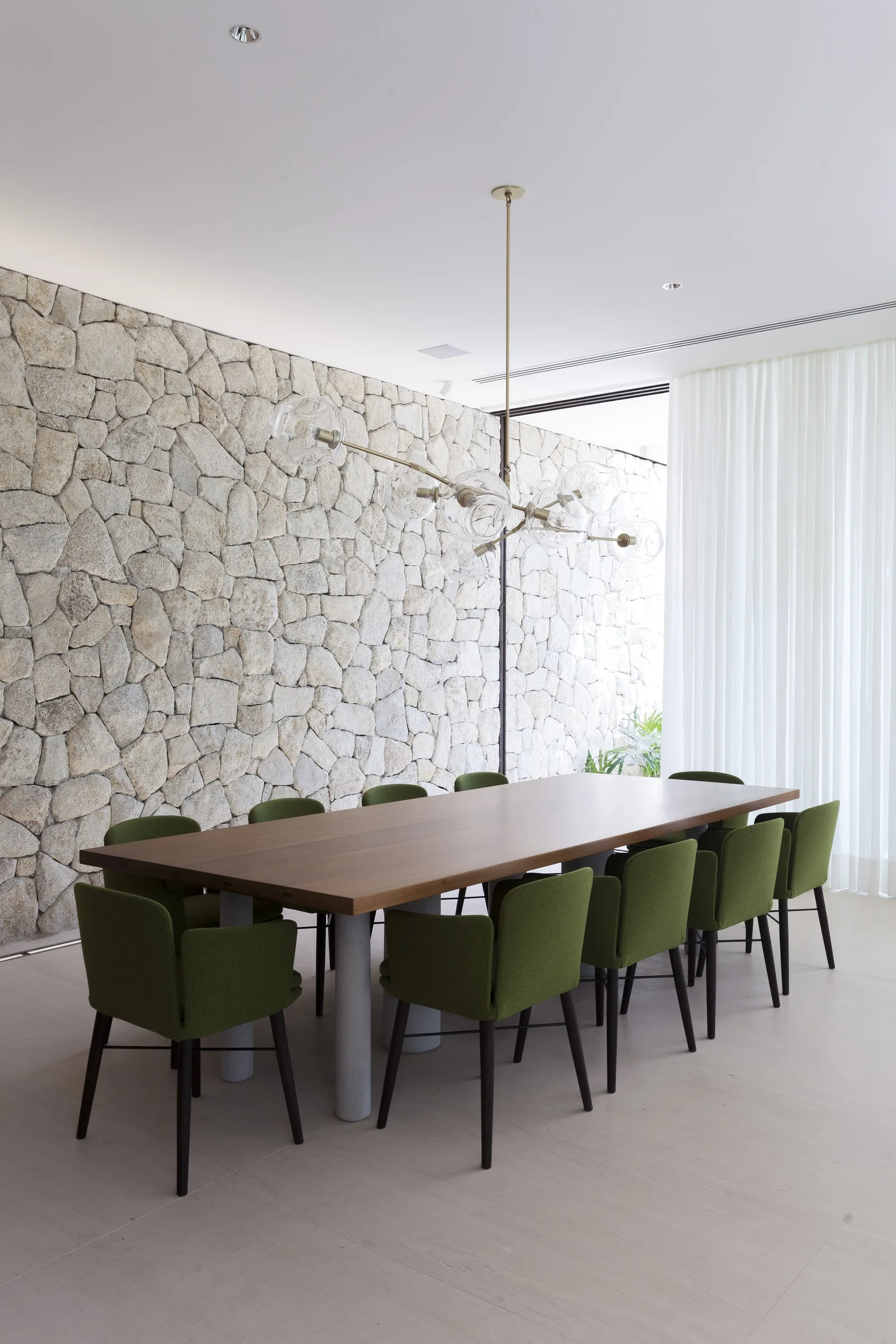 A modern dining room features a long wooden table with green chairs next to a large stacked stone accent wall and sheer white curtains.