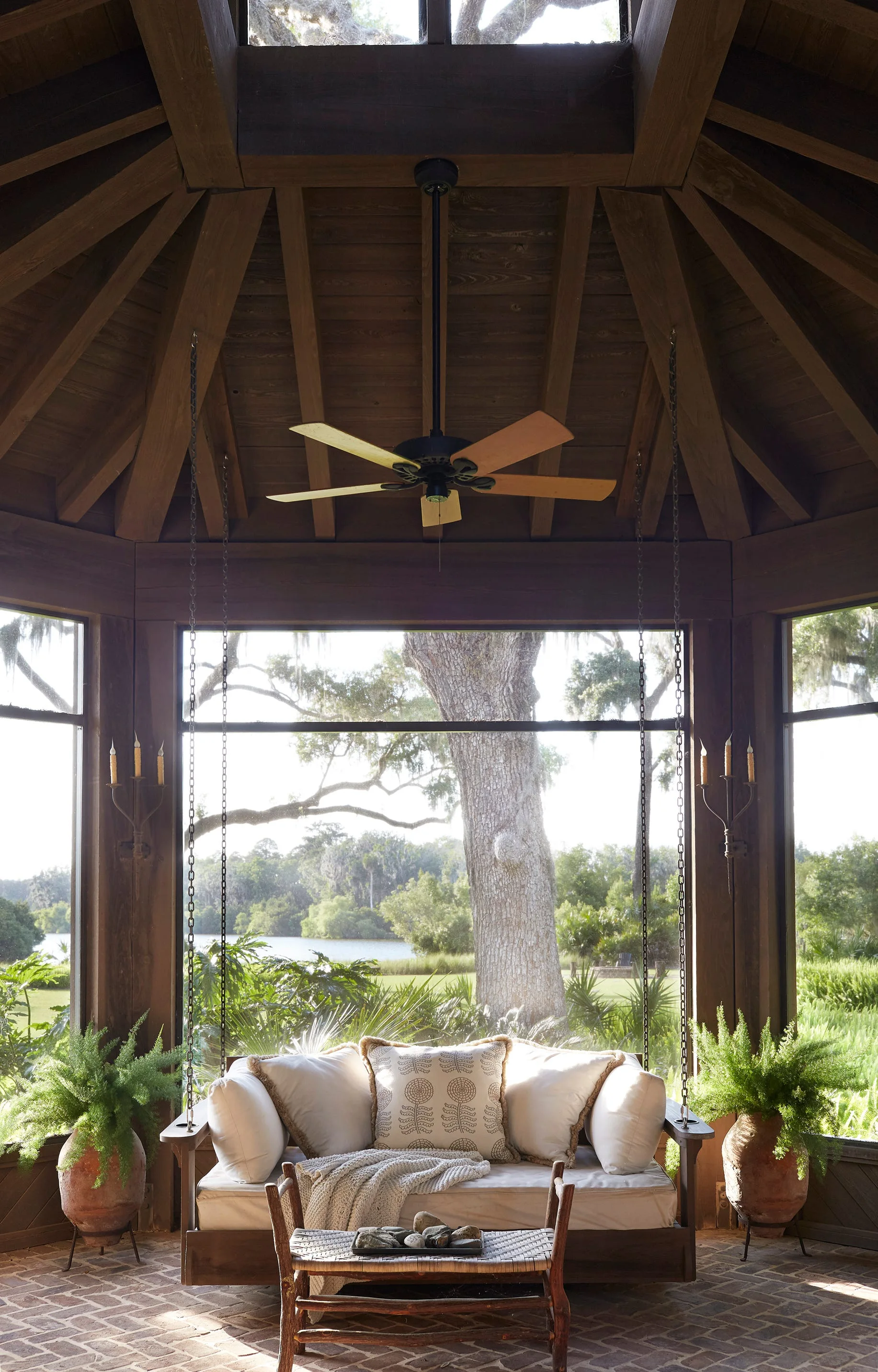 A rustic wooden porch features a suspended daybed swing with white cushions, flanked by two large clay planters with ferns, under a vaulted ceiling.