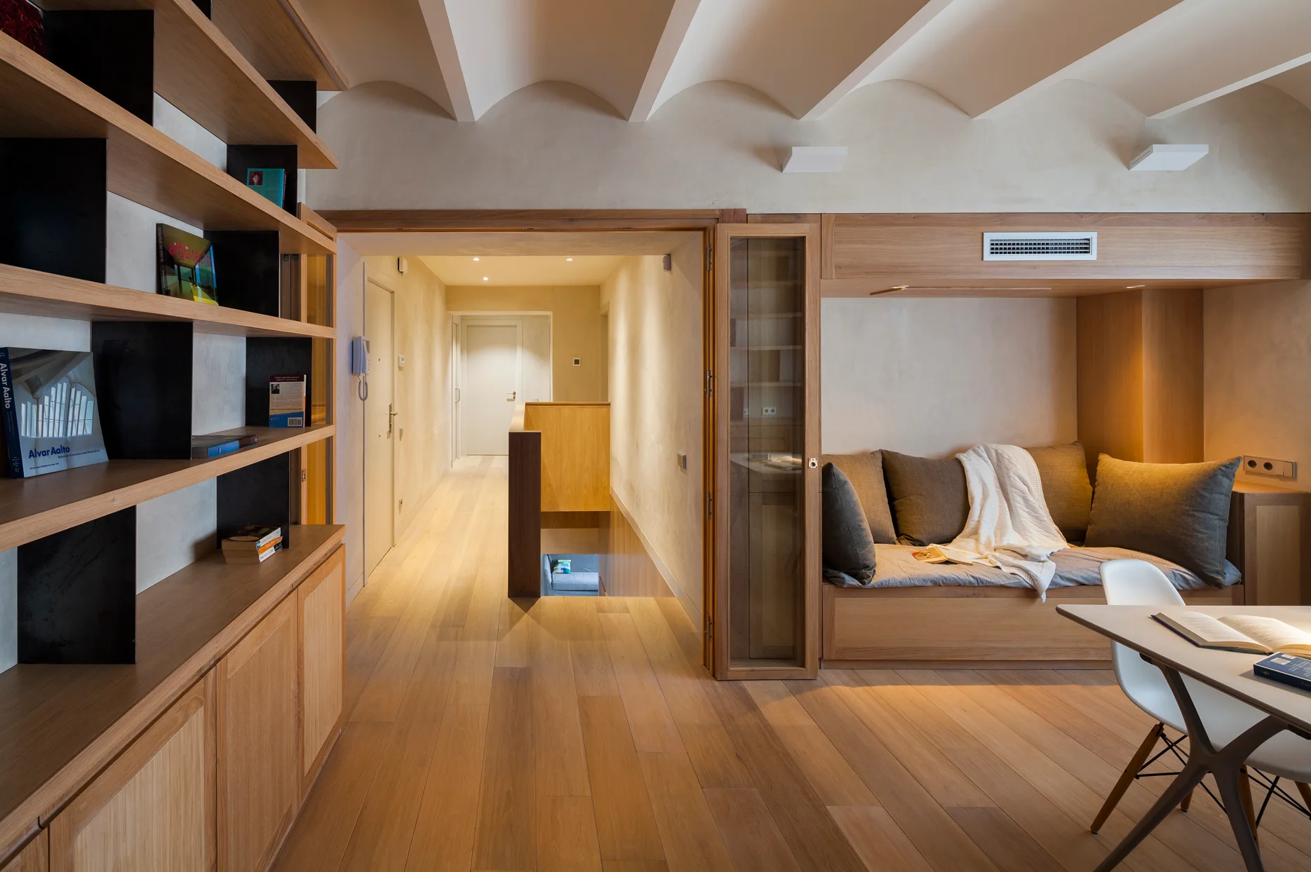 A modern room featuring light wood flooring, a built-in daybed niche, custom wooden bookshelves, and a vaulted ceiling looking toward a hallway.