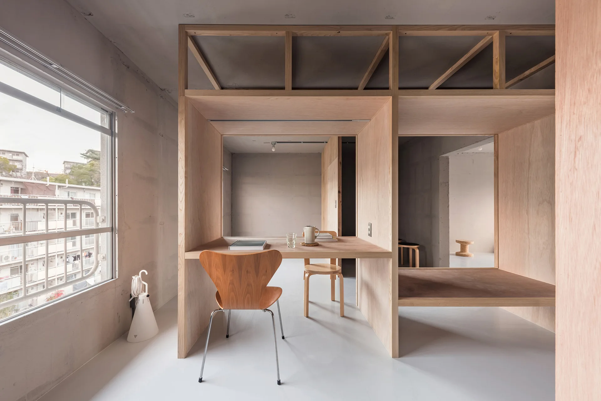 A built-in plywood desk and partition structure sits inside a minimalist room with raw concrete walls, a large window, and a wooden chair.