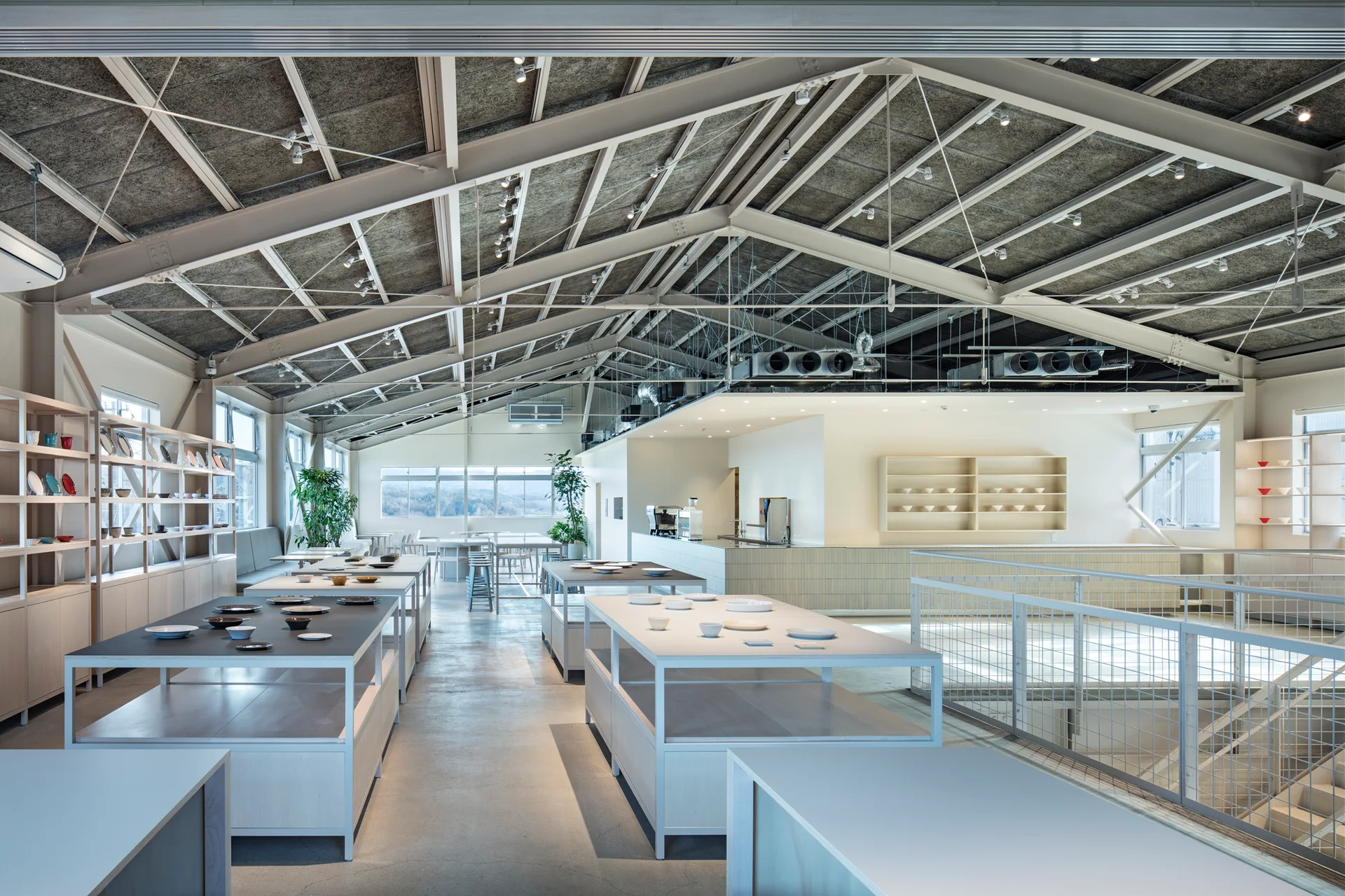 A spacious minimalist showroom features exposed white steel trusses, light grey concrete floors, and light wood display tables with ceramic plates.