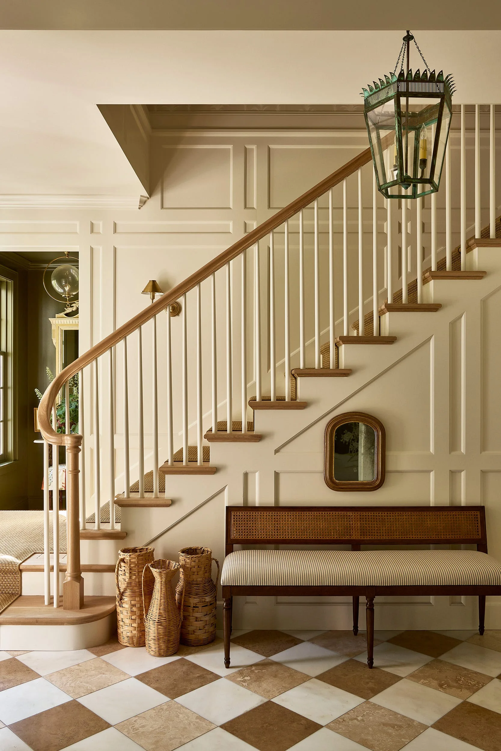 A cream-colored foyer features a wooden staircase, a striped upholstered bench with a rattan back, and diagonal checkerboard stone floor tiles.