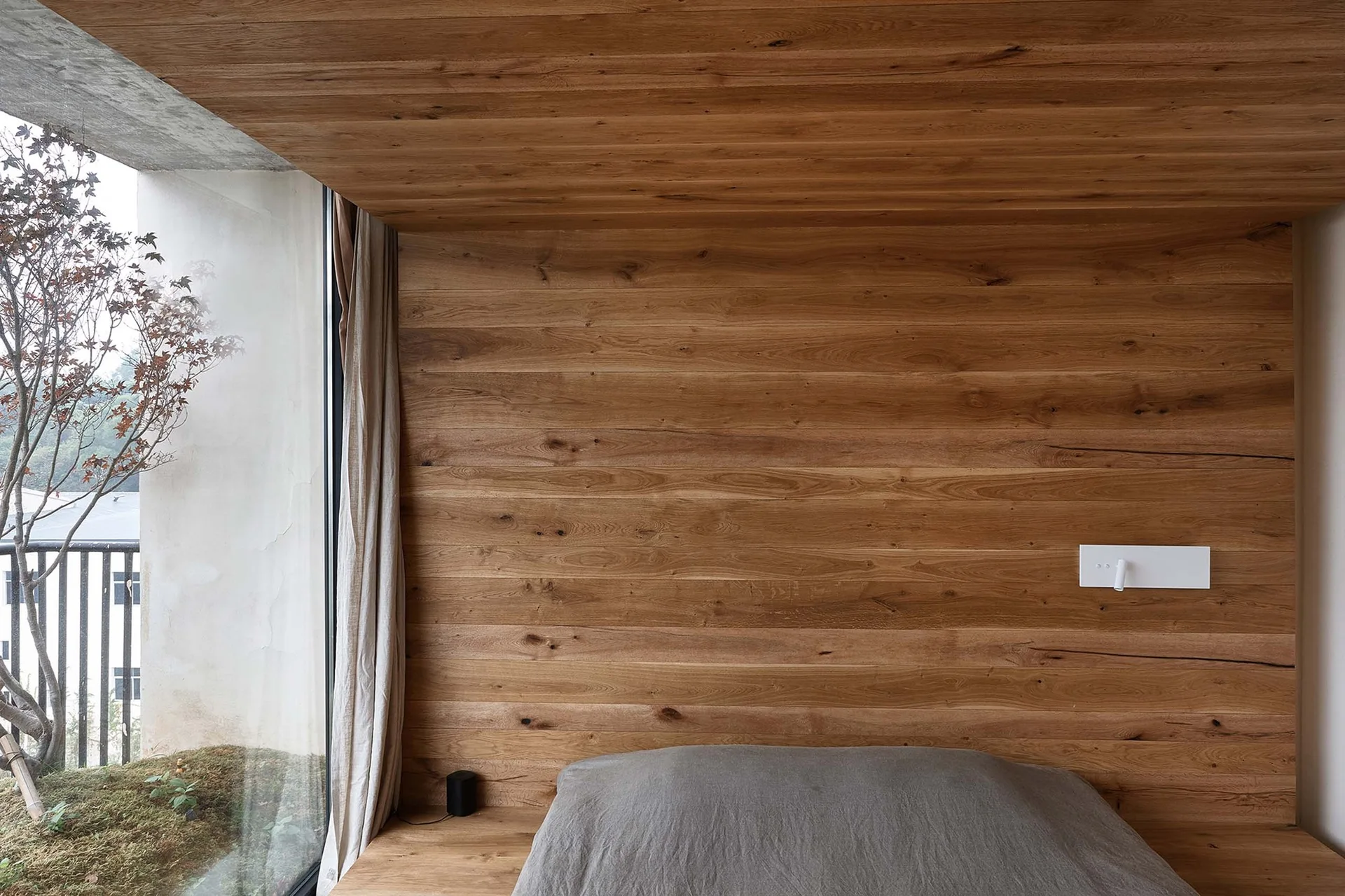 A minimalist bedroom featuring horizontal wood planking on the walls and ceiling, a platform bed with grey linens, and a large window.