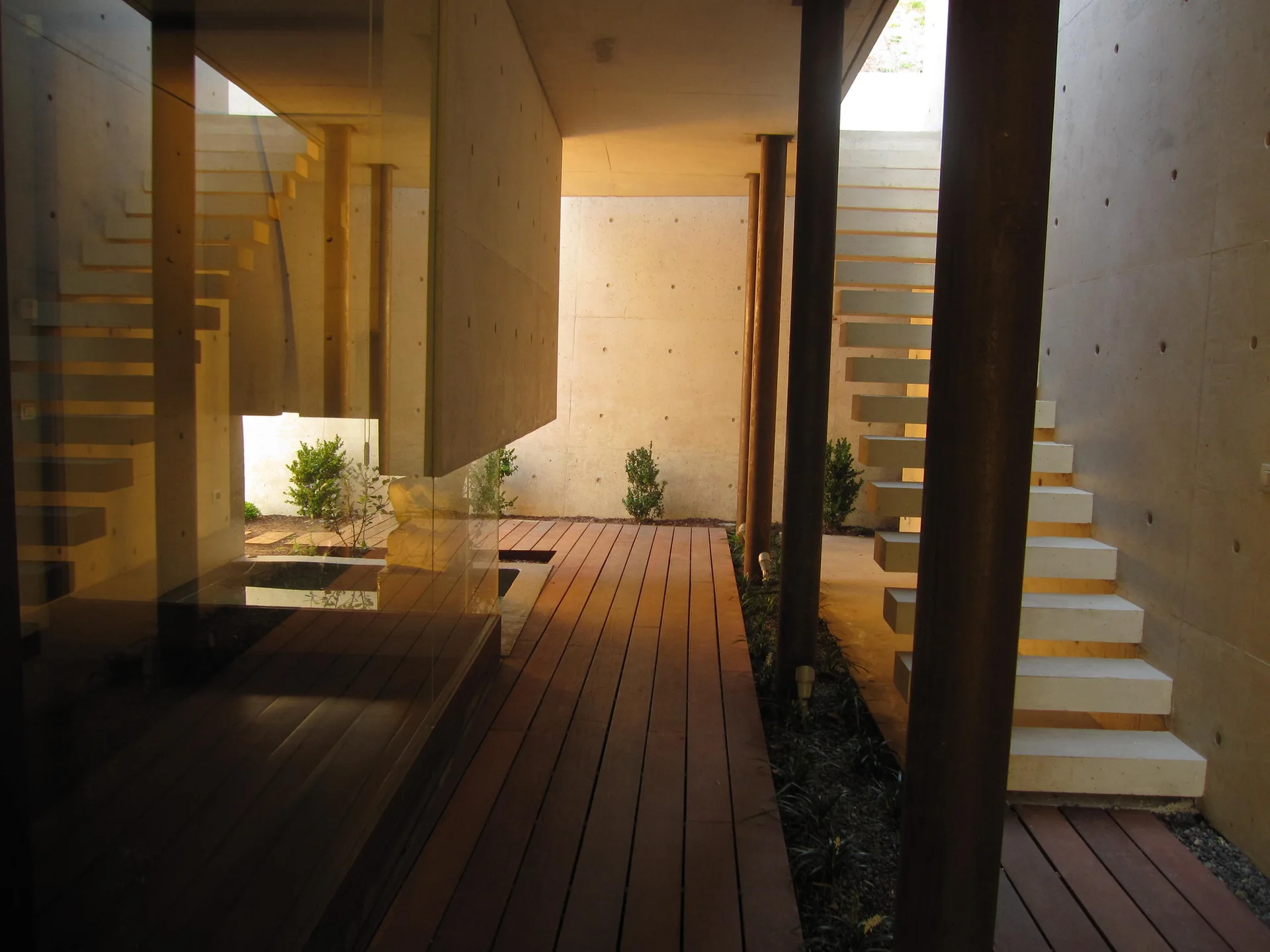 A modern outdoor courtyard with a wood plank deck, raw concrete walls, structural columns, and a floating concrete staircase.