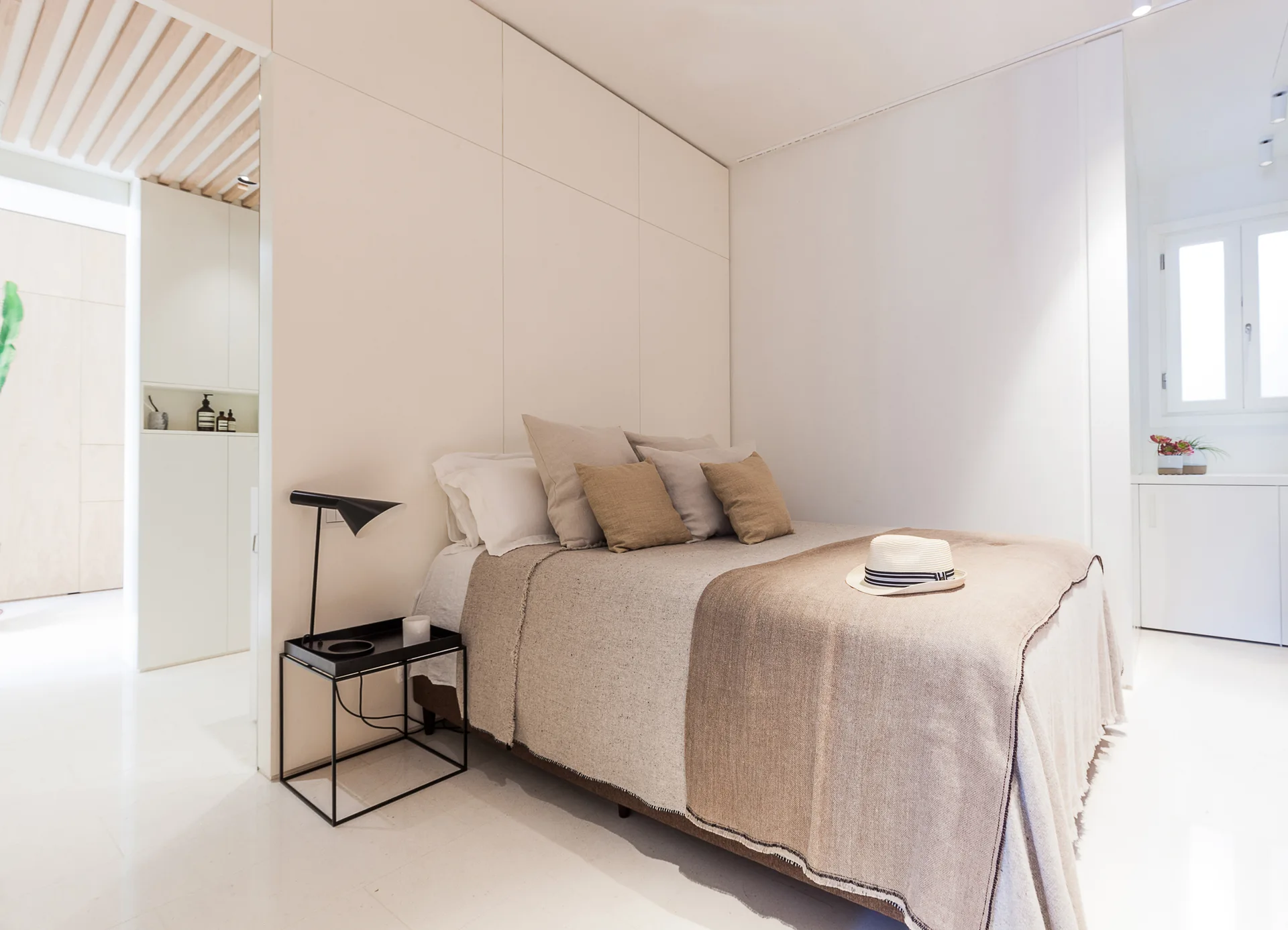 A minimalist bedroom featuring a bed with beige linens set against a white paneled wall beside a black metal nightstand and lamp.