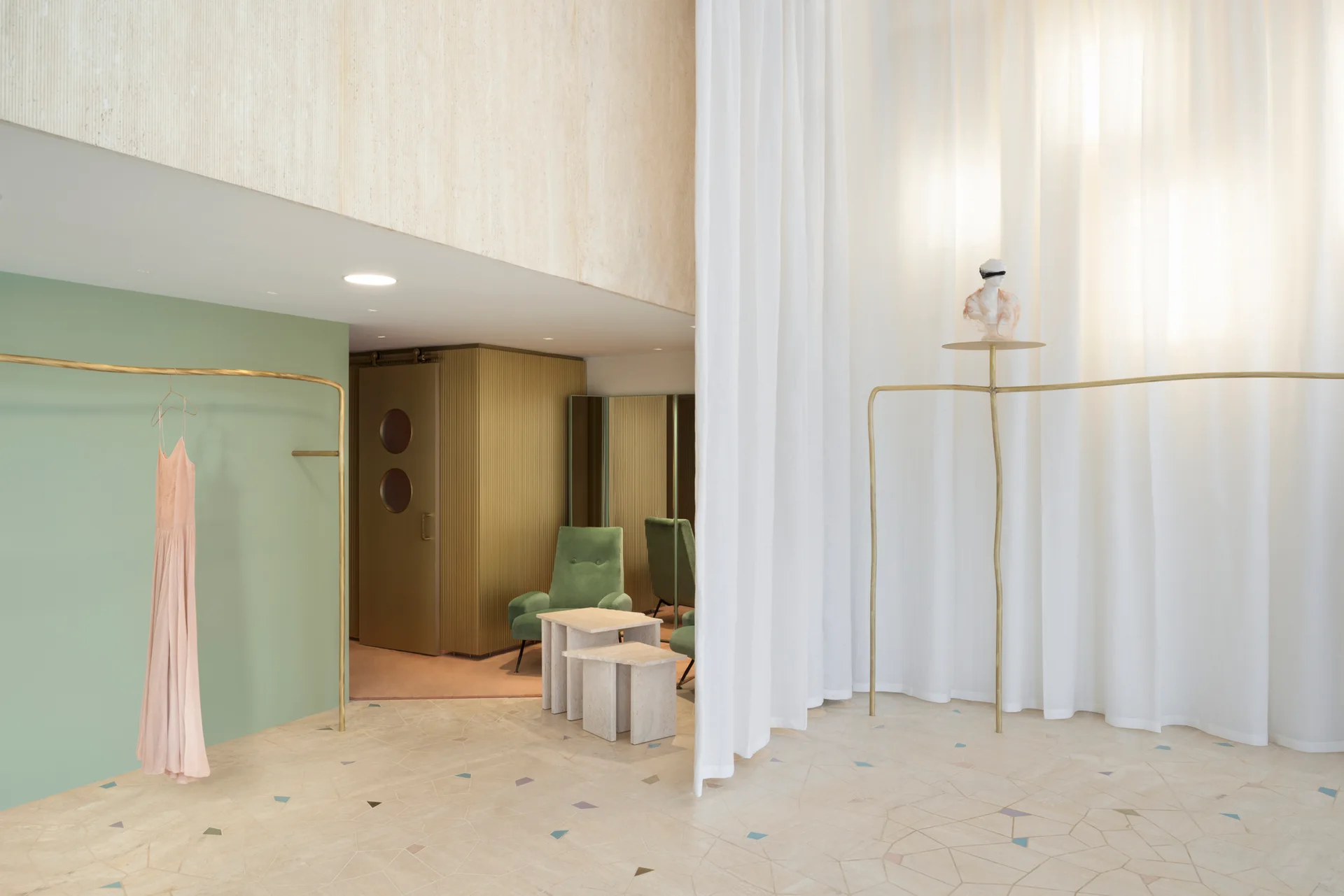 A minimalist retail interior features curved brass clothing racks, a sage green wall, sheer white drapes, and stone floor tiles with geometric inlays.