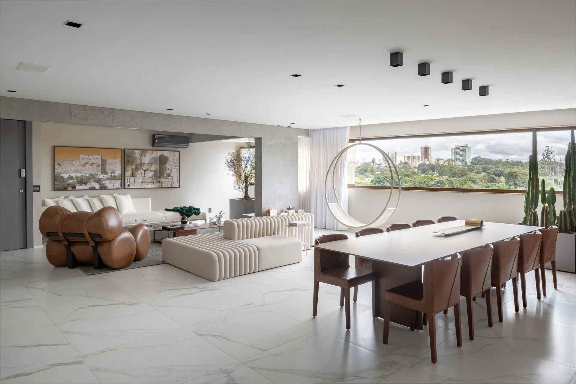 A spacious open-plan living and dining room with a ribbed beige modular sofa, a sculptural leather armchair, and a long wooden dining table on marble floors.