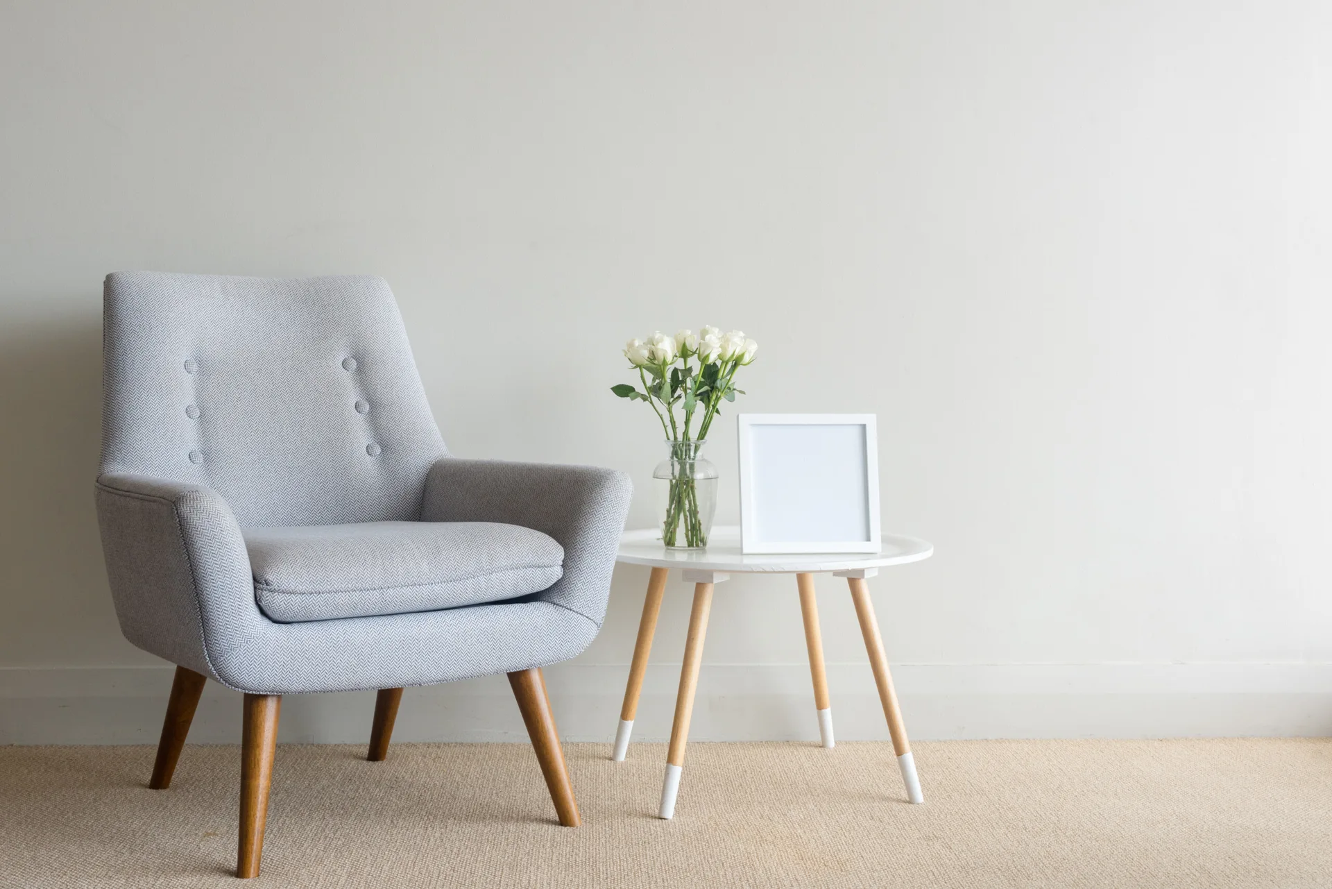 A light grey tufted armchair with wooden legs sits next to a small white side table holding white roses and a picture frame against a plain wall.