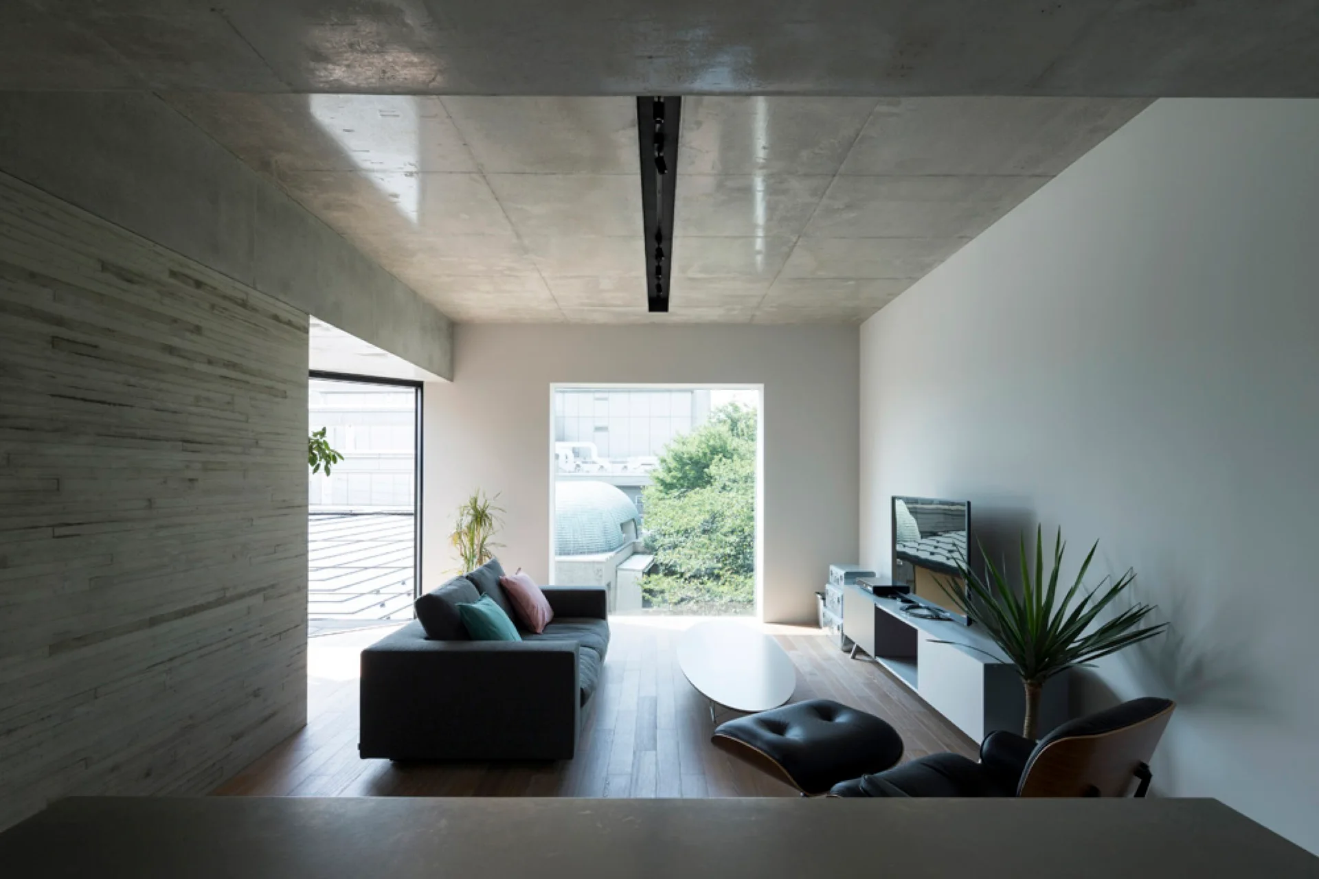 A minimalist living room featuring a dark gray sofa, a leather lounge chair, and a large window under an exposed concrete ceiling.