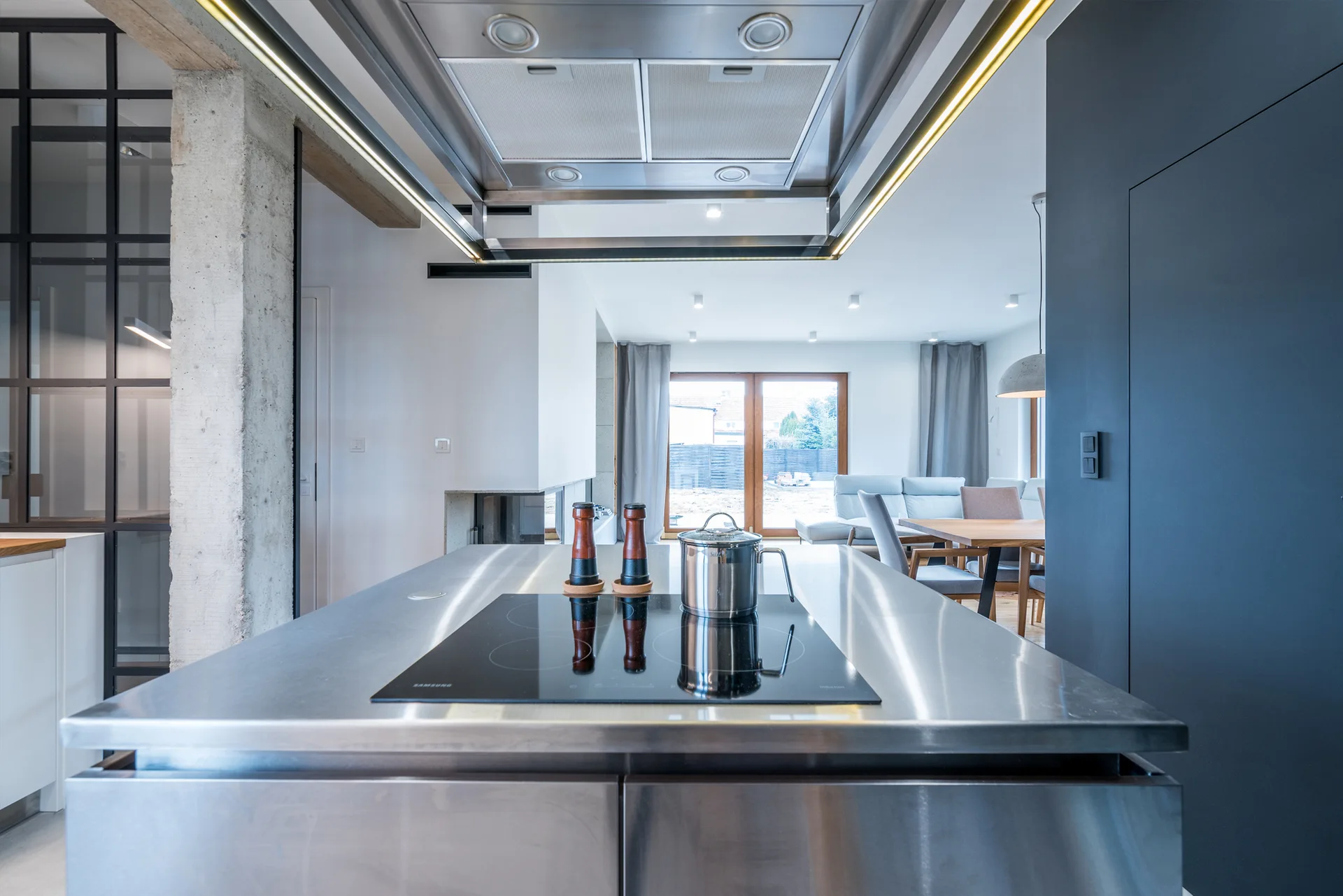 A modern open-concept kitchen features a large stainless steel island with a black cooktop, an exposed concrete column, and a dining area in the background.