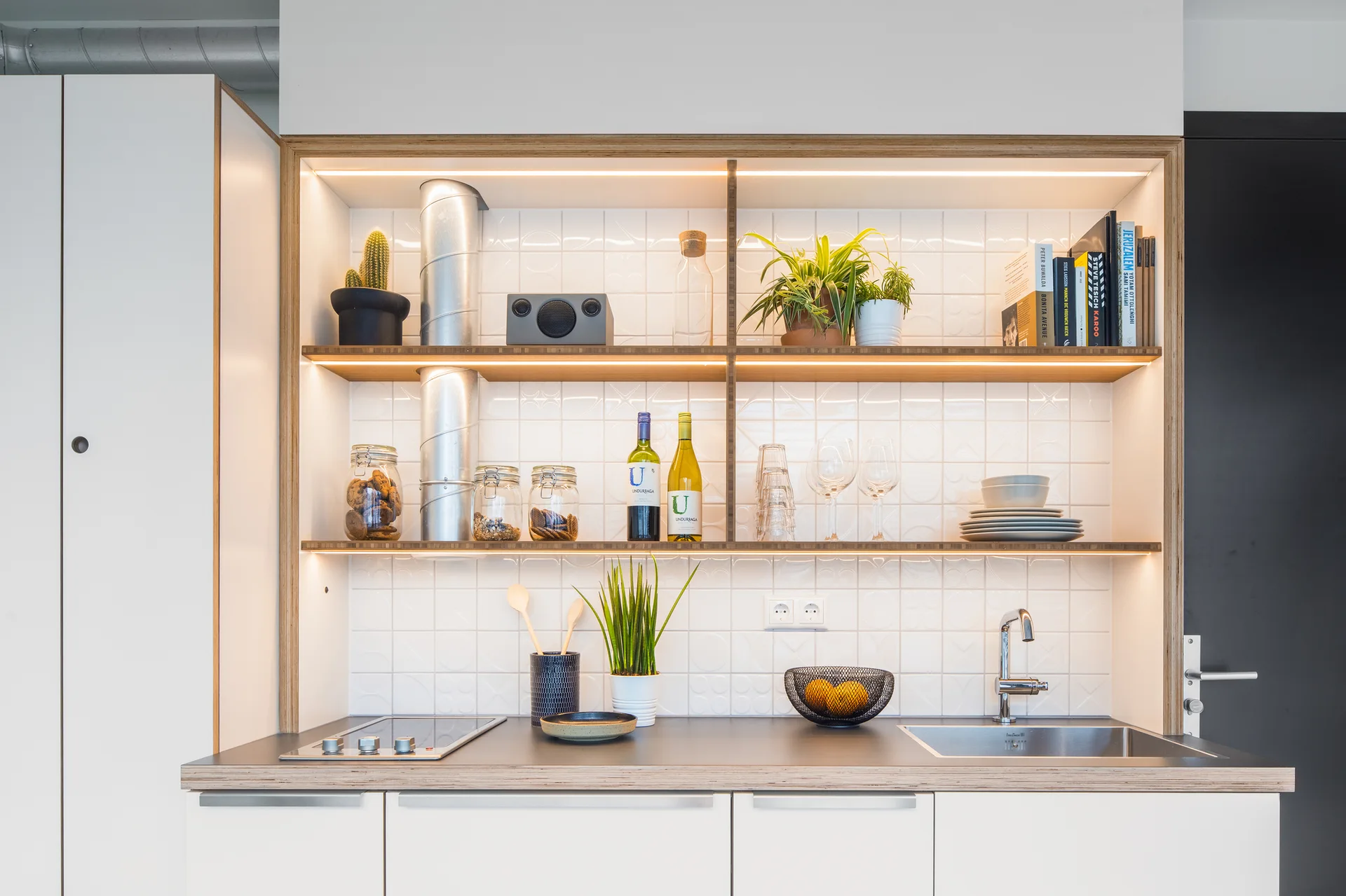 A compact kitchen with white cabinetry, a square-tiled backsplash, and built-in plywood shelving illuminated by warm LED lighting.