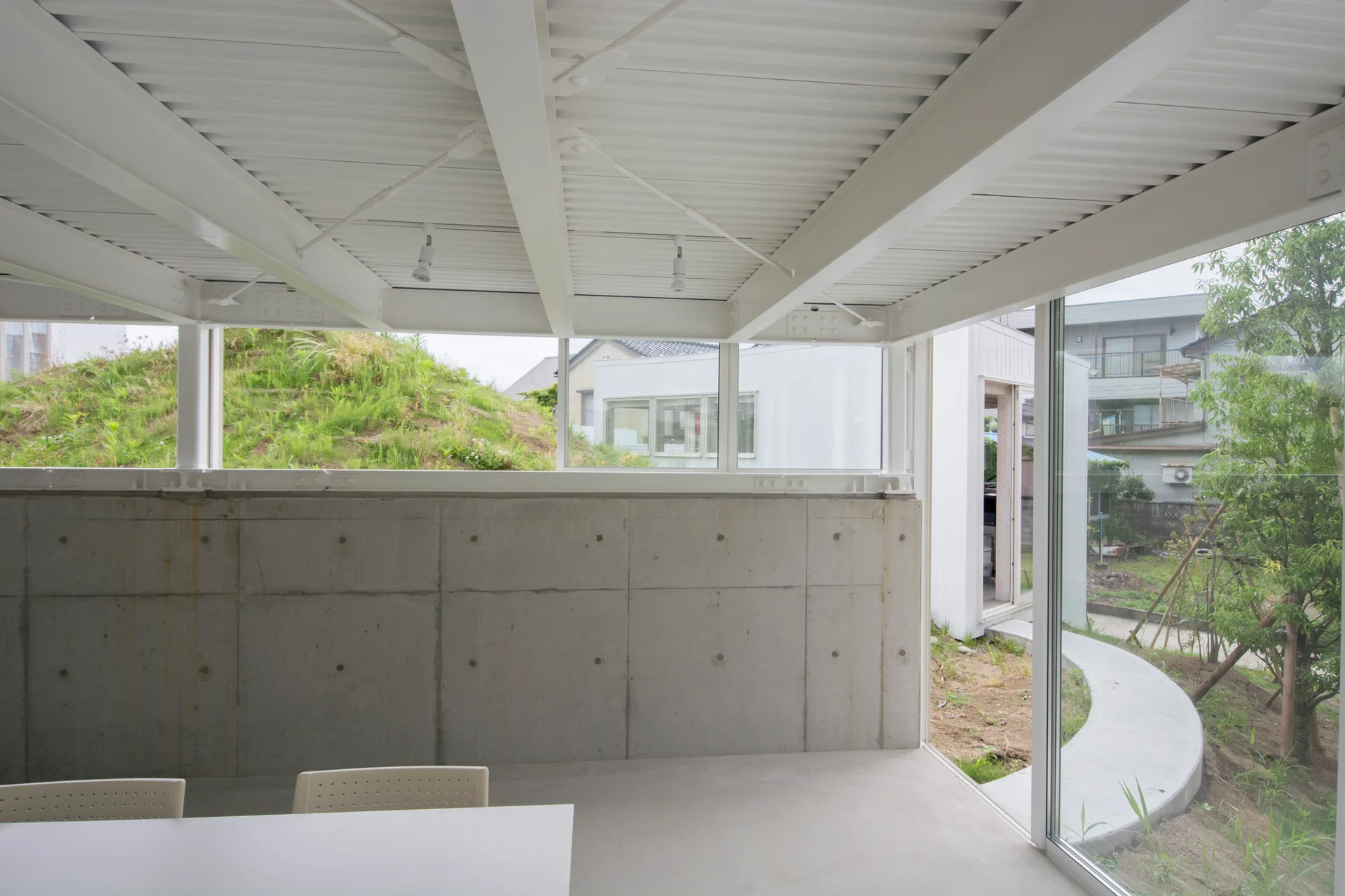 A minimalist room featuring exposed concrete walls, a white structural steel ceiling, and a large glass window looking out to a grassy hill.