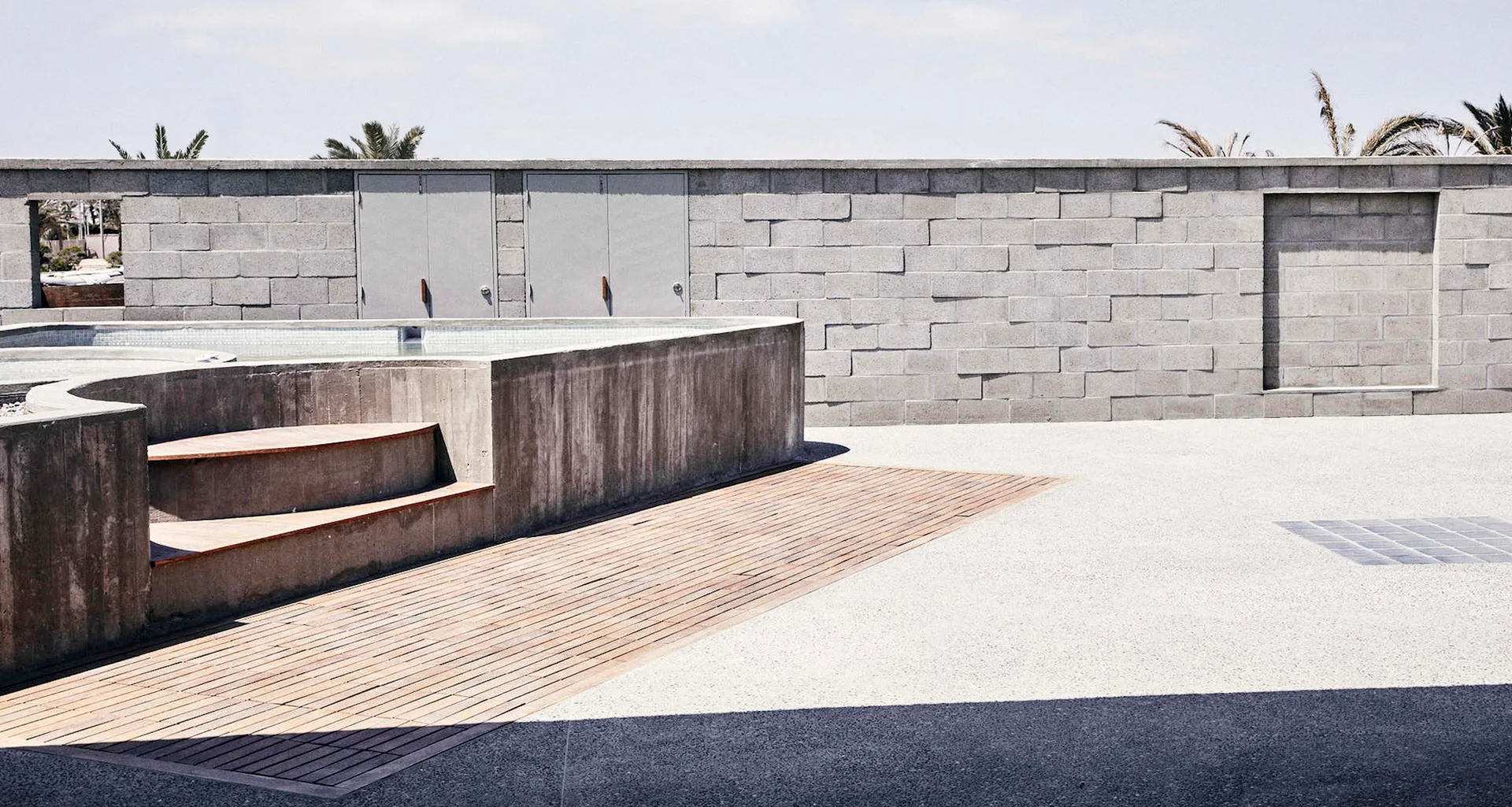 A minimalist outdoor pool area features a poured concrete structure with inset wooden steps and a matching wood plank deck against a gray block wall.