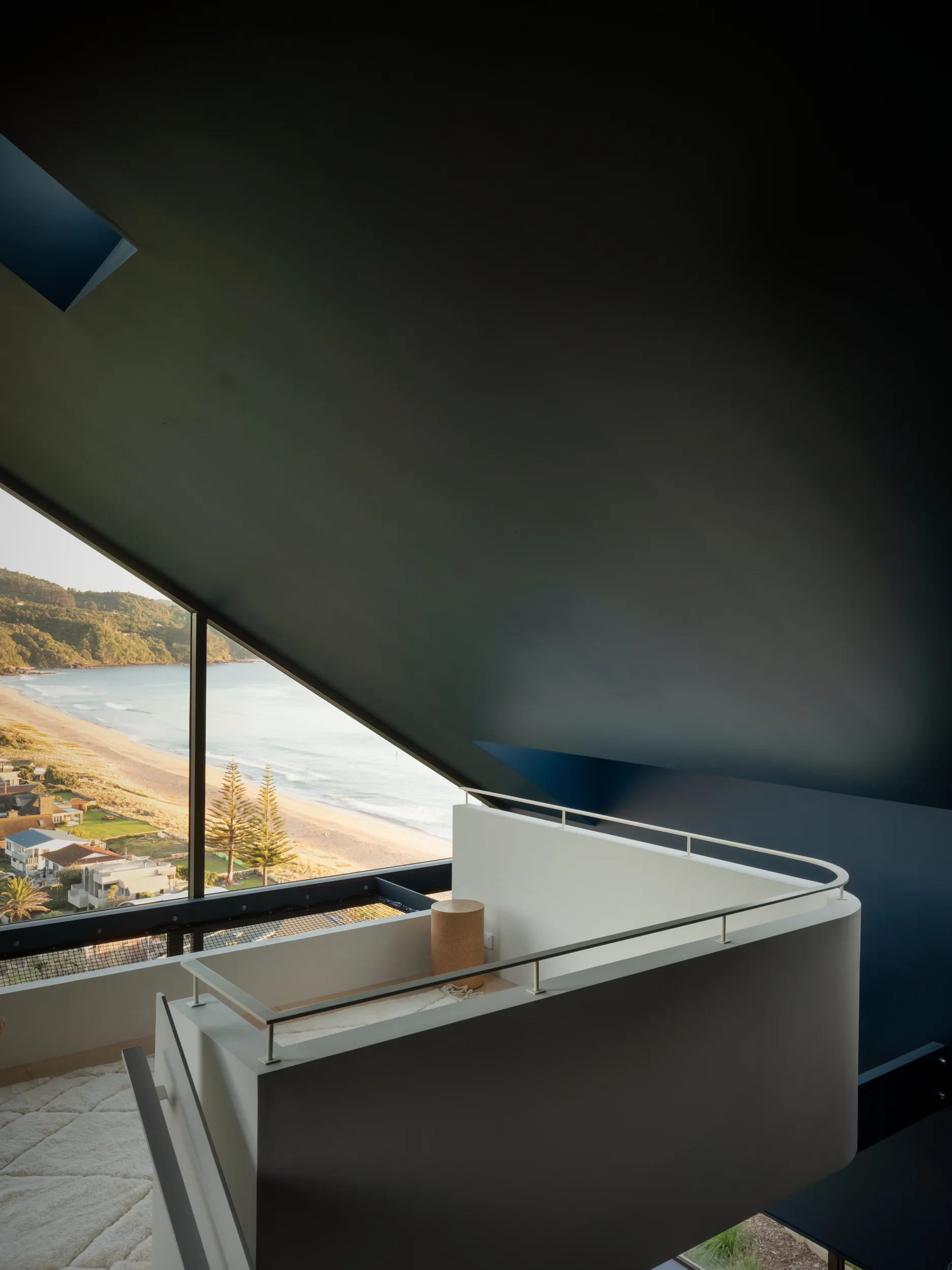 A modern mezzanine landing features a curved white balustrade and a small cork stool beneath a slanted dark blue ceiling with a scenic coastal window view.