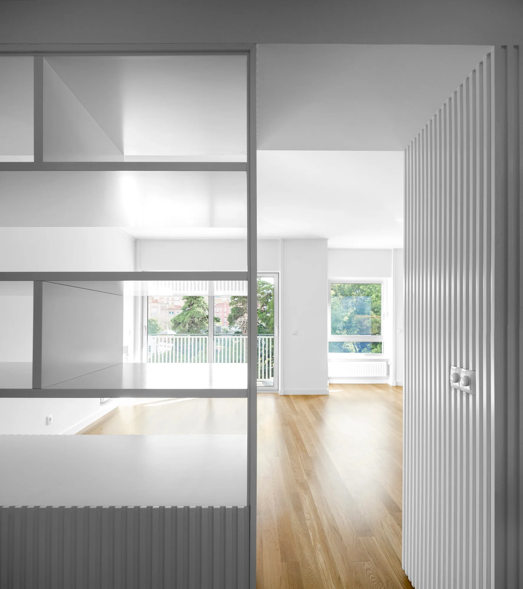 A view through a white open-shelving room divider showing a bright living area with light hardwood floors and vertical ribbed wall paneling.
