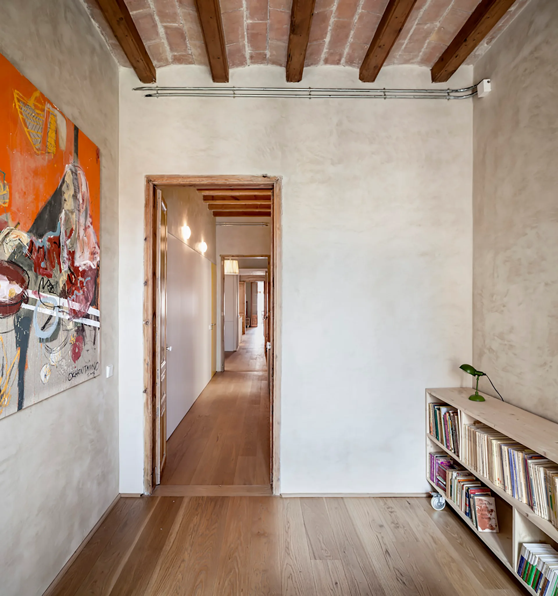 A long residential hallway featuring textured plaster walls, exposed wood ceiling beams with brick, and a low wooden bookcase on wheels.