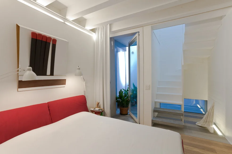 A minimalist white bedroom features a bed with a bright red headboard, white exposed ceiling beams, and a modern graphic print of matches on the wall.