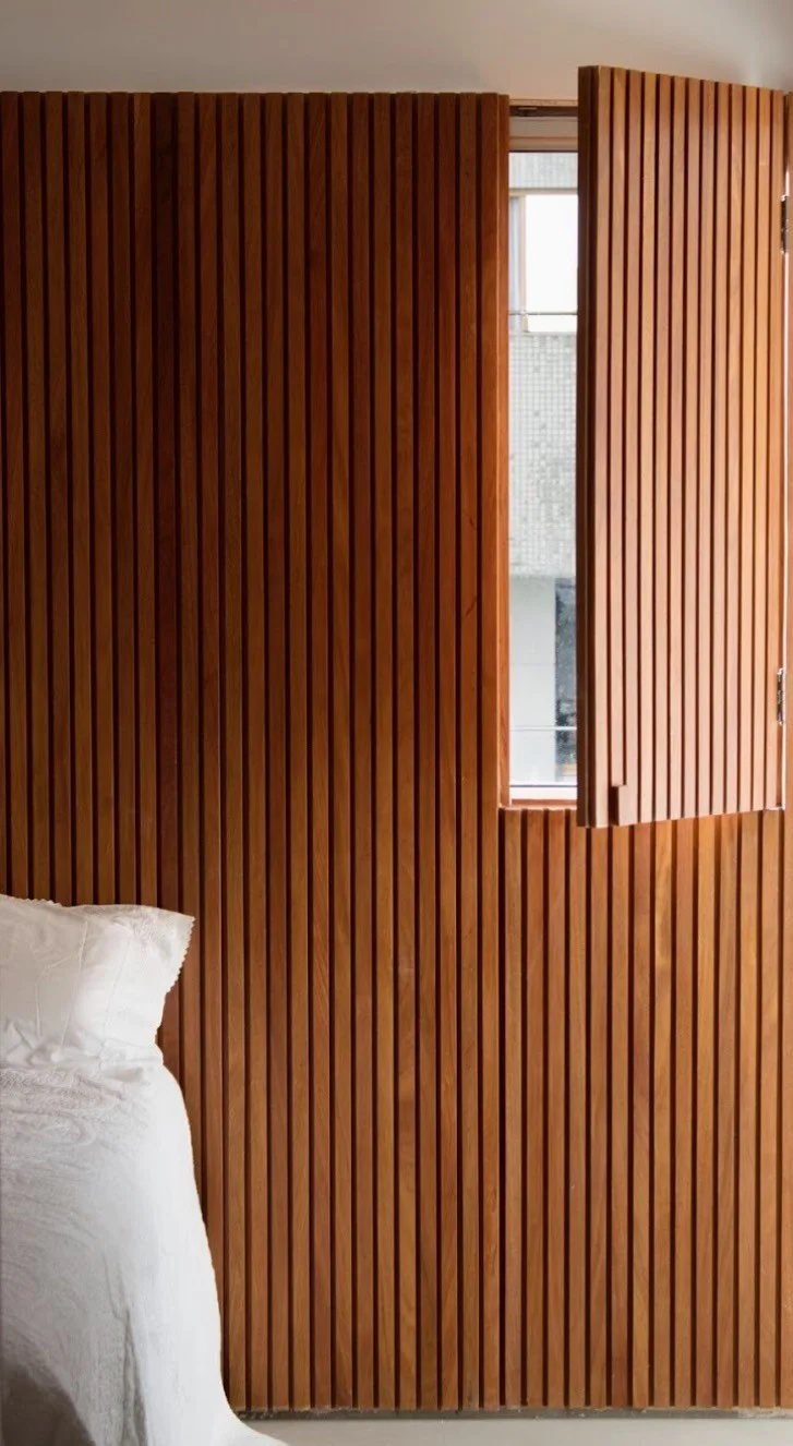 A bedroom corner shows warm wood vertical slat wall paneling and a matching hinged shutter opened to reveal a small window next to a white bed.