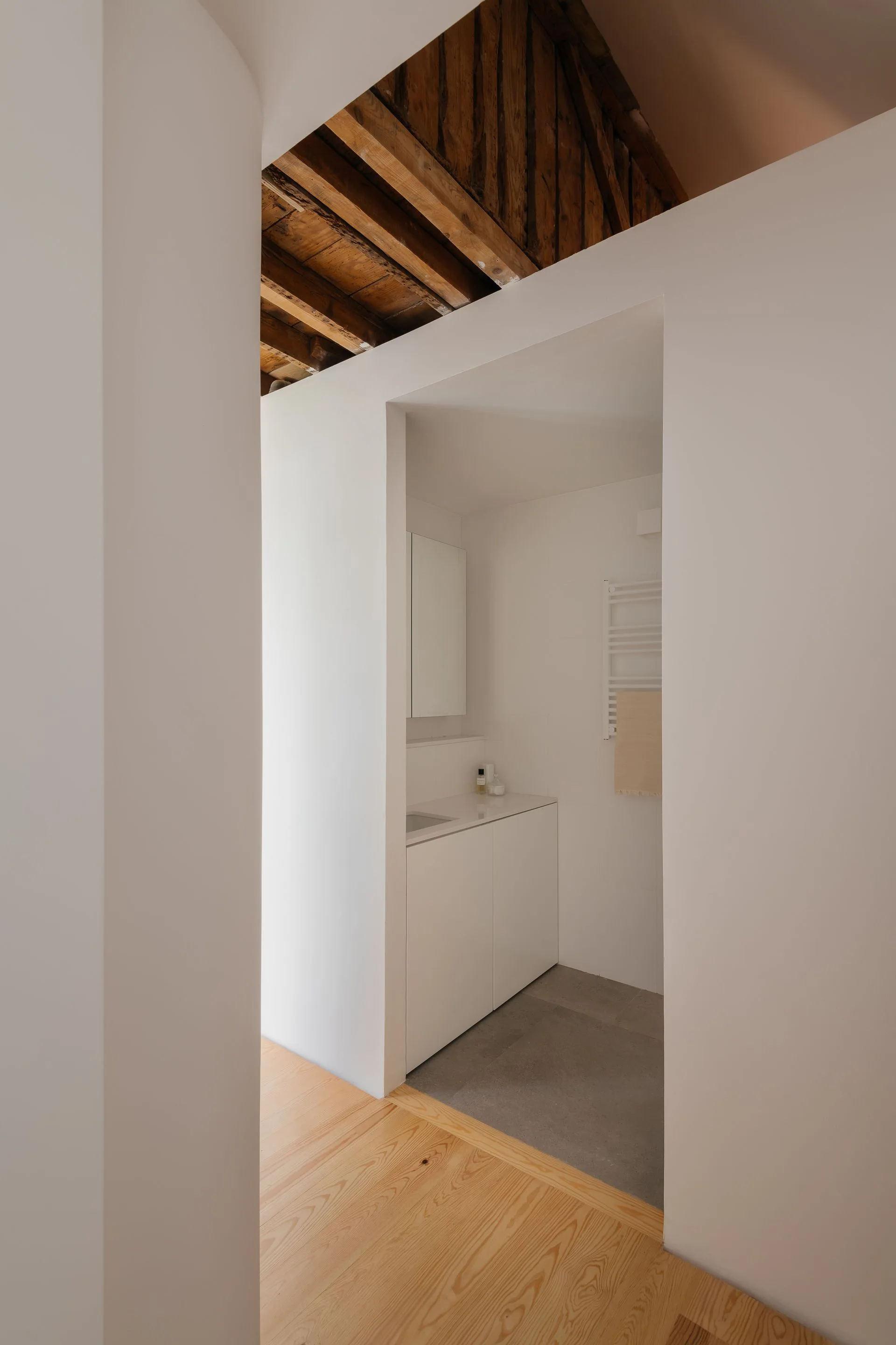 A minimalist white bathroom with a built-in vanity and towel radiator under an exposed rustic wood ceiling, viewed through a recessed entryway.