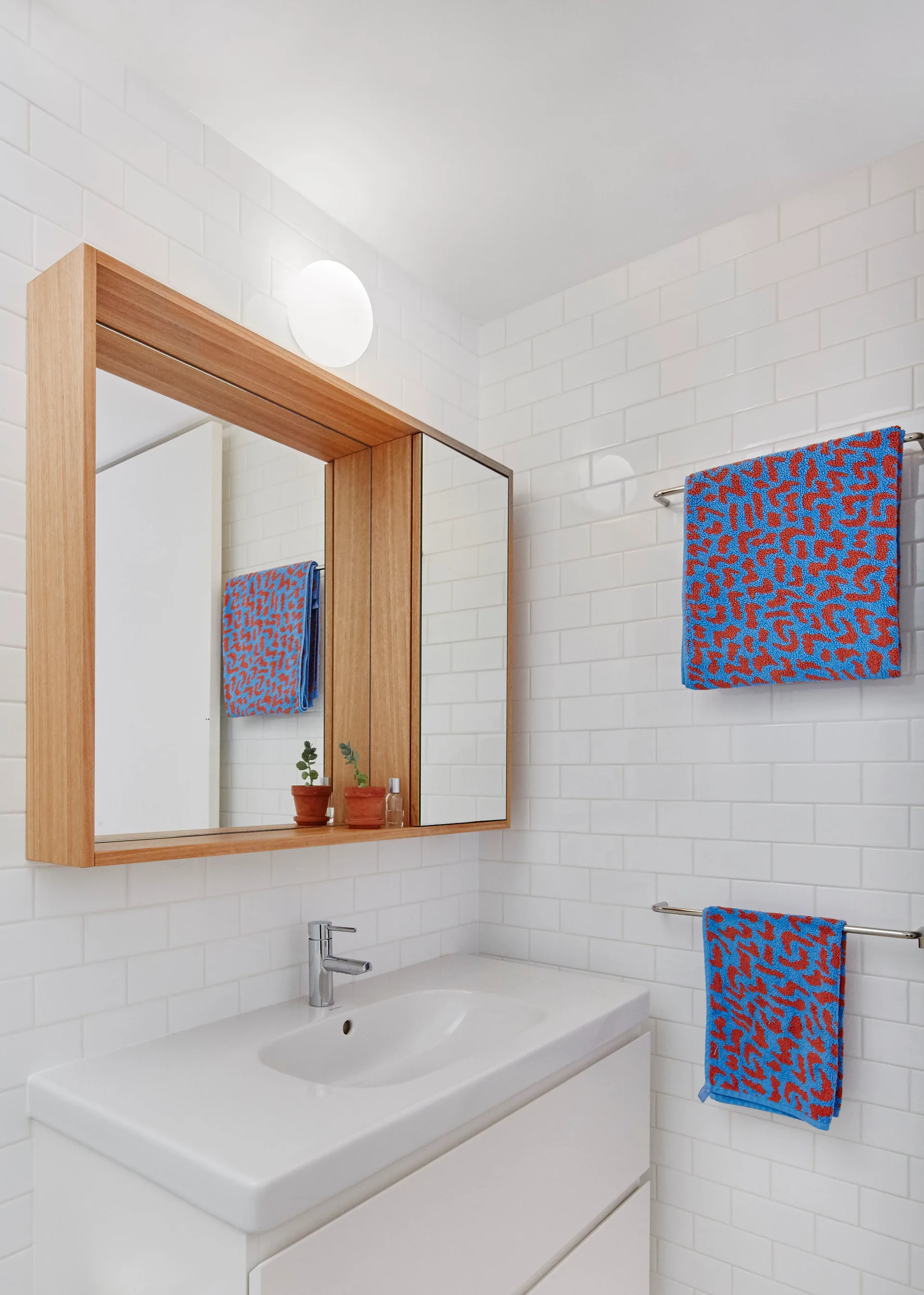 A modern white bathroom vanity with a wood-framed medicine cabinet on a subway tile wall next to patterned blue and red towels.