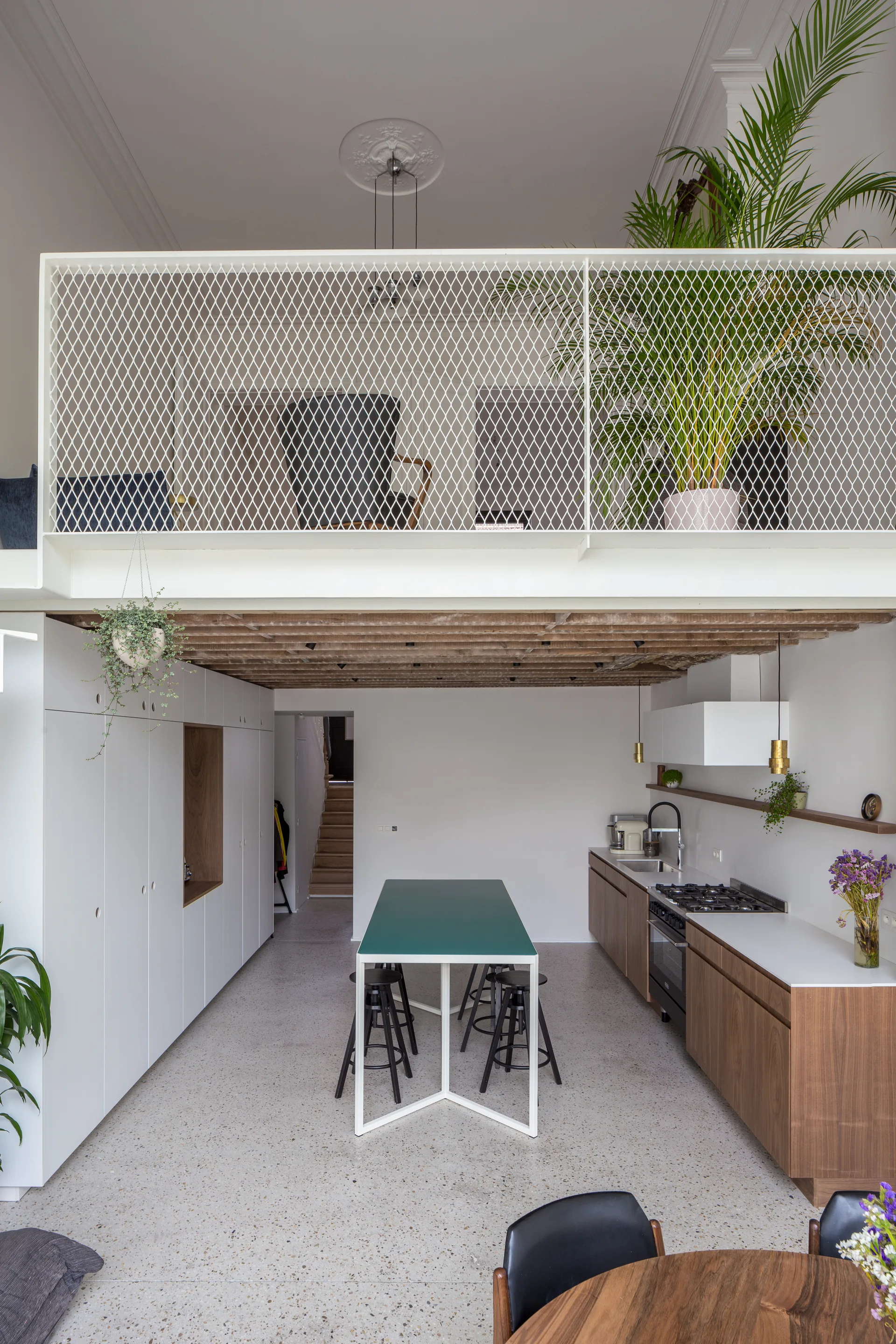 A modern kitchen features a green-topped island, wooden cabinetry, and a mezzanine level with a white metal mesh railing.