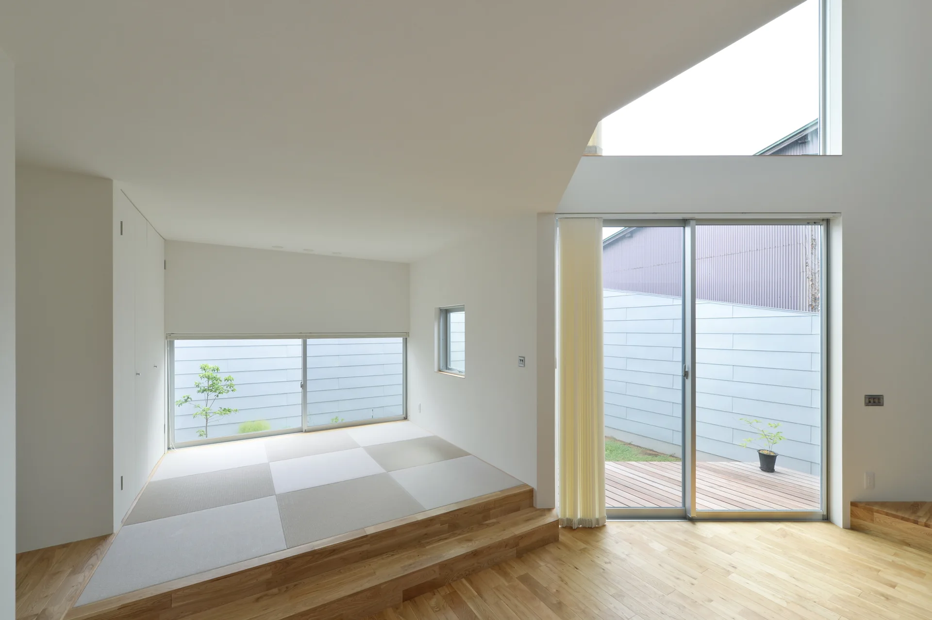 A minimalist white room features a raised wooden platform with tatami mats, light hardwood floors, and sliding glass doors leading to a deck.