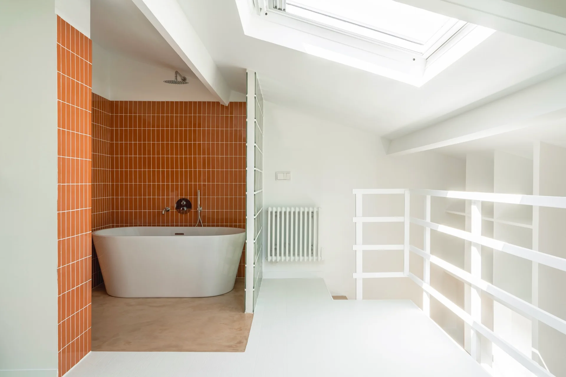 A modern attic bathroom featuring a white freestanding bathtub against an orange tiled wall next to a glass block partition.