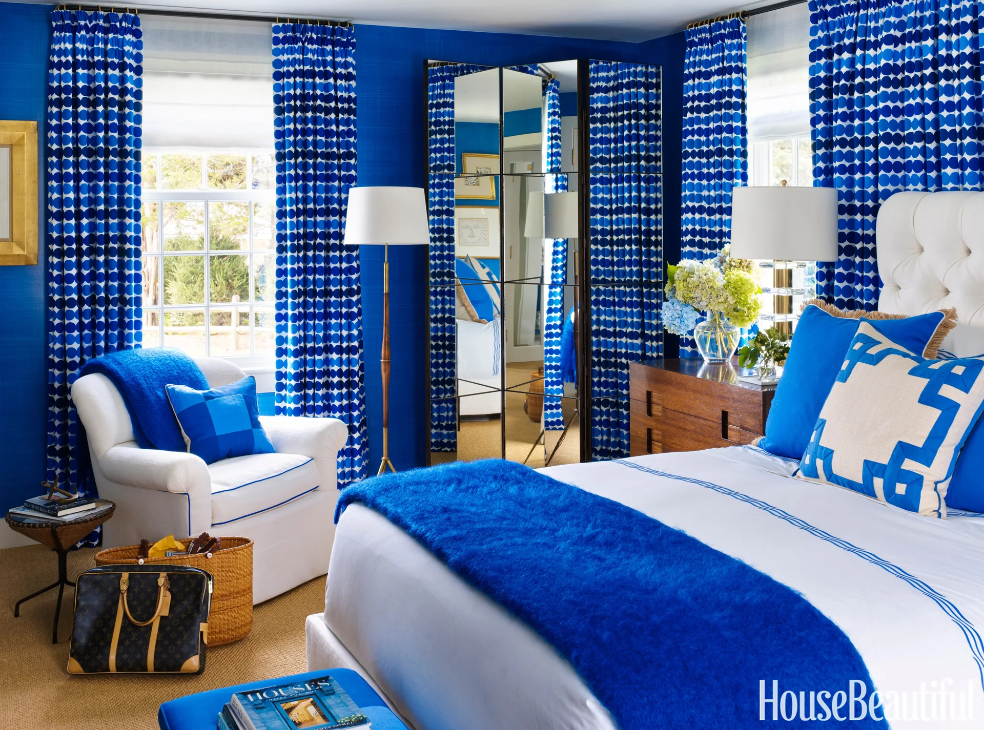 A bedroom with textured cobalt blue walls, patterned blue and white curtains, a white armchair, and a tall mirrored folding screen.