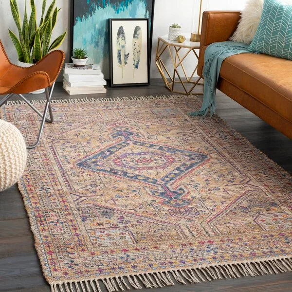 A brown leather sofa, tan butterfly chair, and geometric gold side table sit on a large multicolored tasseled rug on dark wood floors.