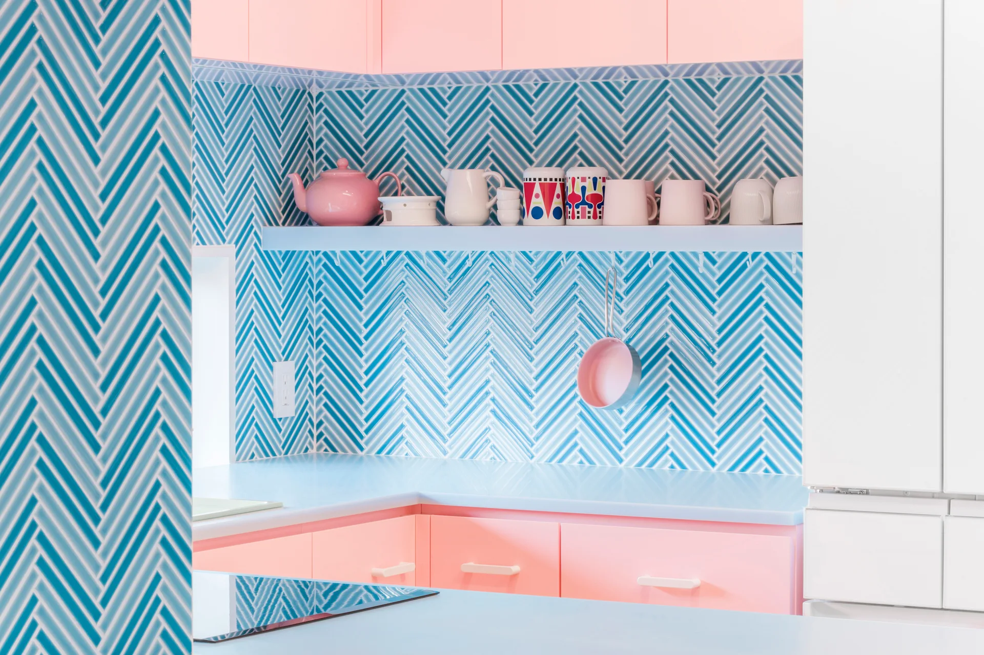 A bright kitchen features pastel pink cabinets beneath a blue-and-white herringbone tile backsplash and a light blue countertop with a floating shelf.