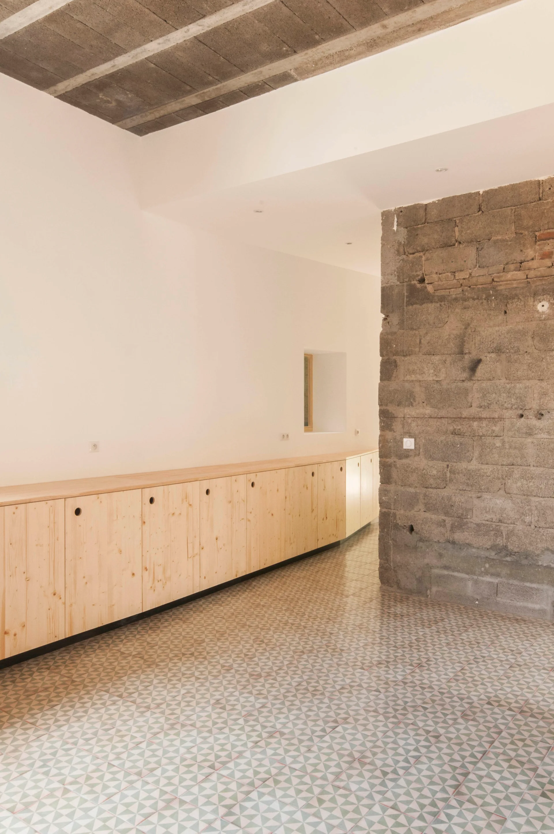 A long row of light wood cabinets sits along a white wall opposite an exposed concrete block pillar on a geometric patterned tile floor.