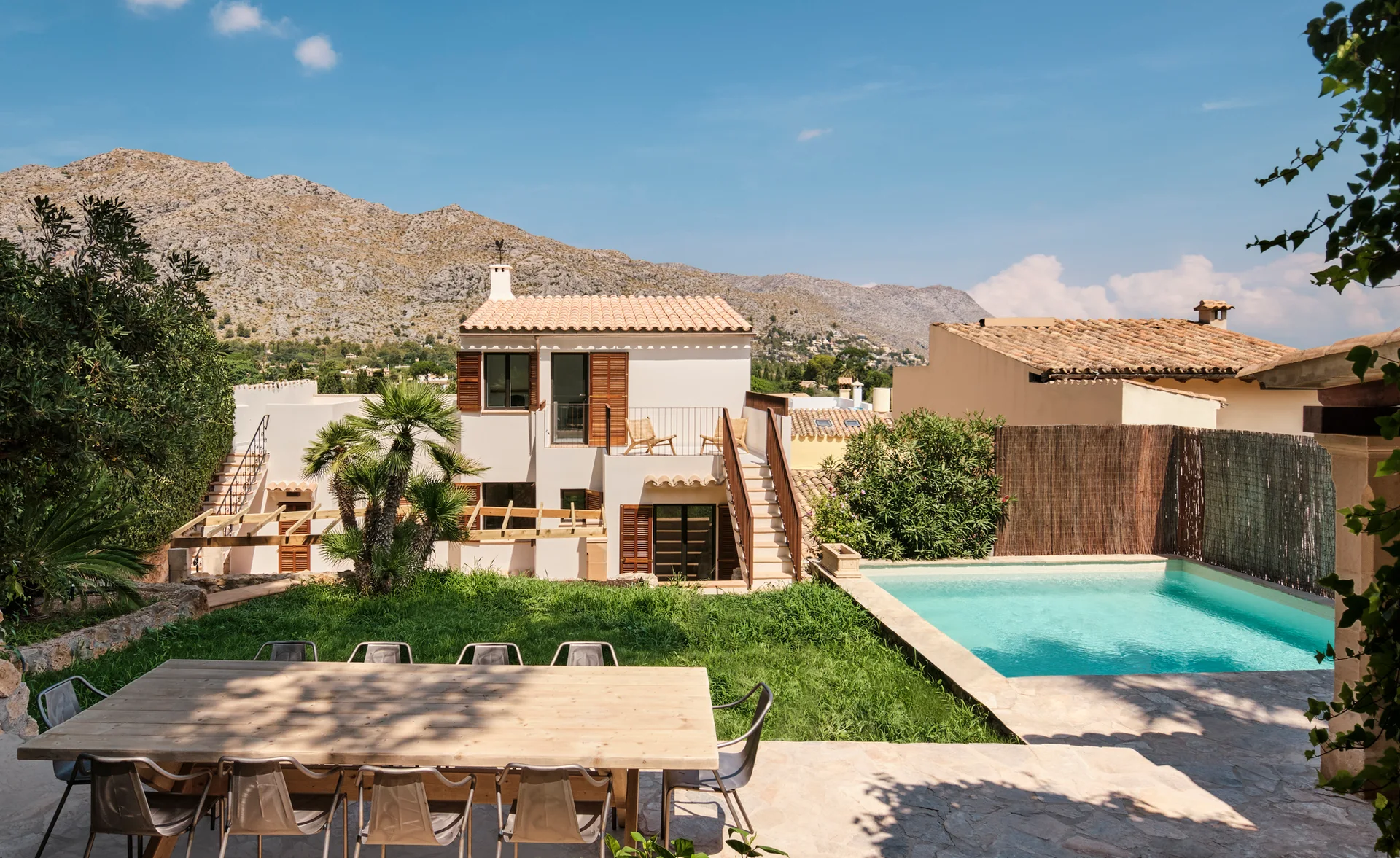 An outdoor stone patio with a wooden dining table overlooks a grassy lawn, a turquoise swimming pool, and a cream stucco villa with mountains behind.
